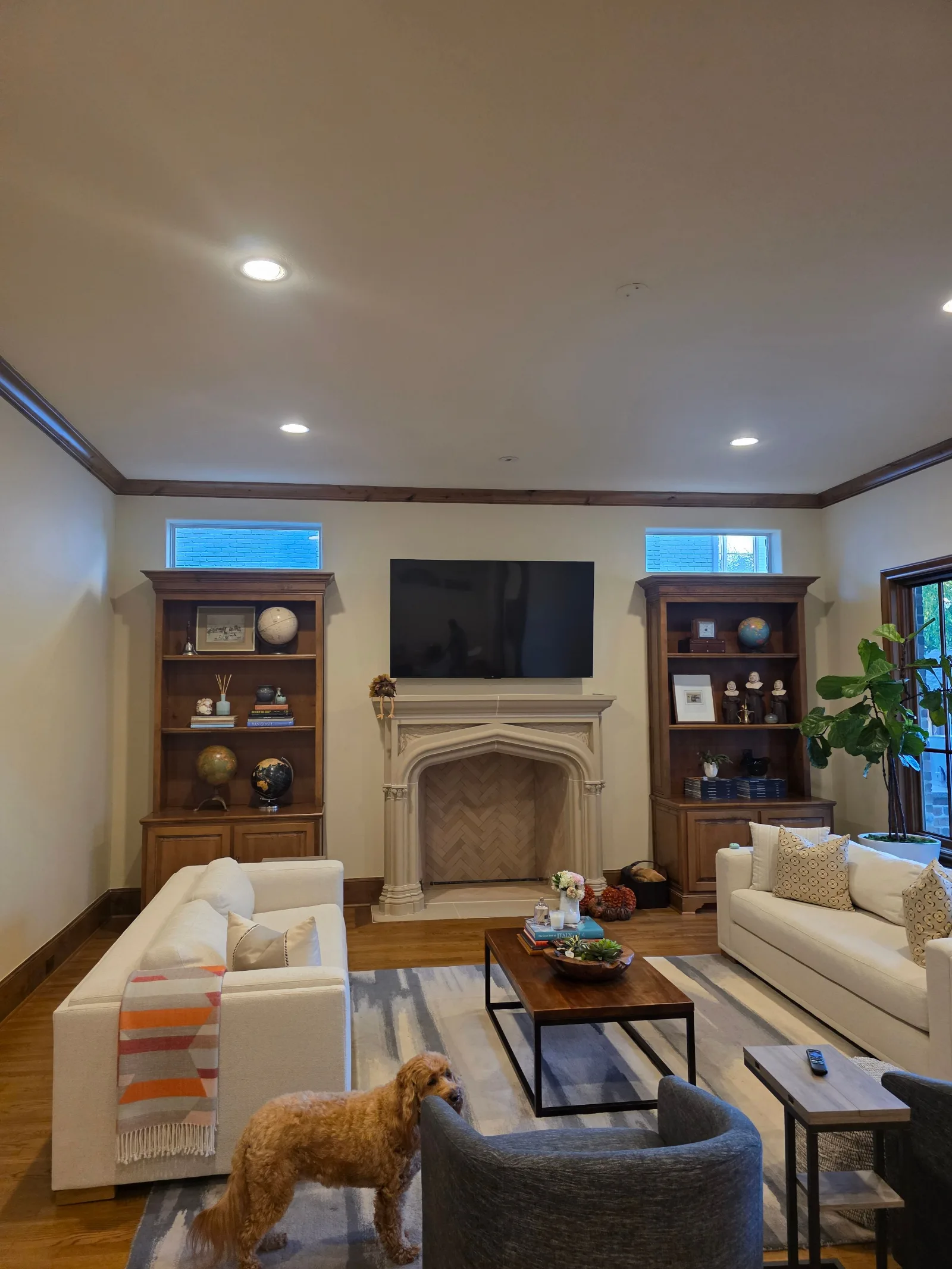 Living room with recessed lighting, brown built-in bookcases, white sofas, wood coffee table, and a carved stone fireplace.