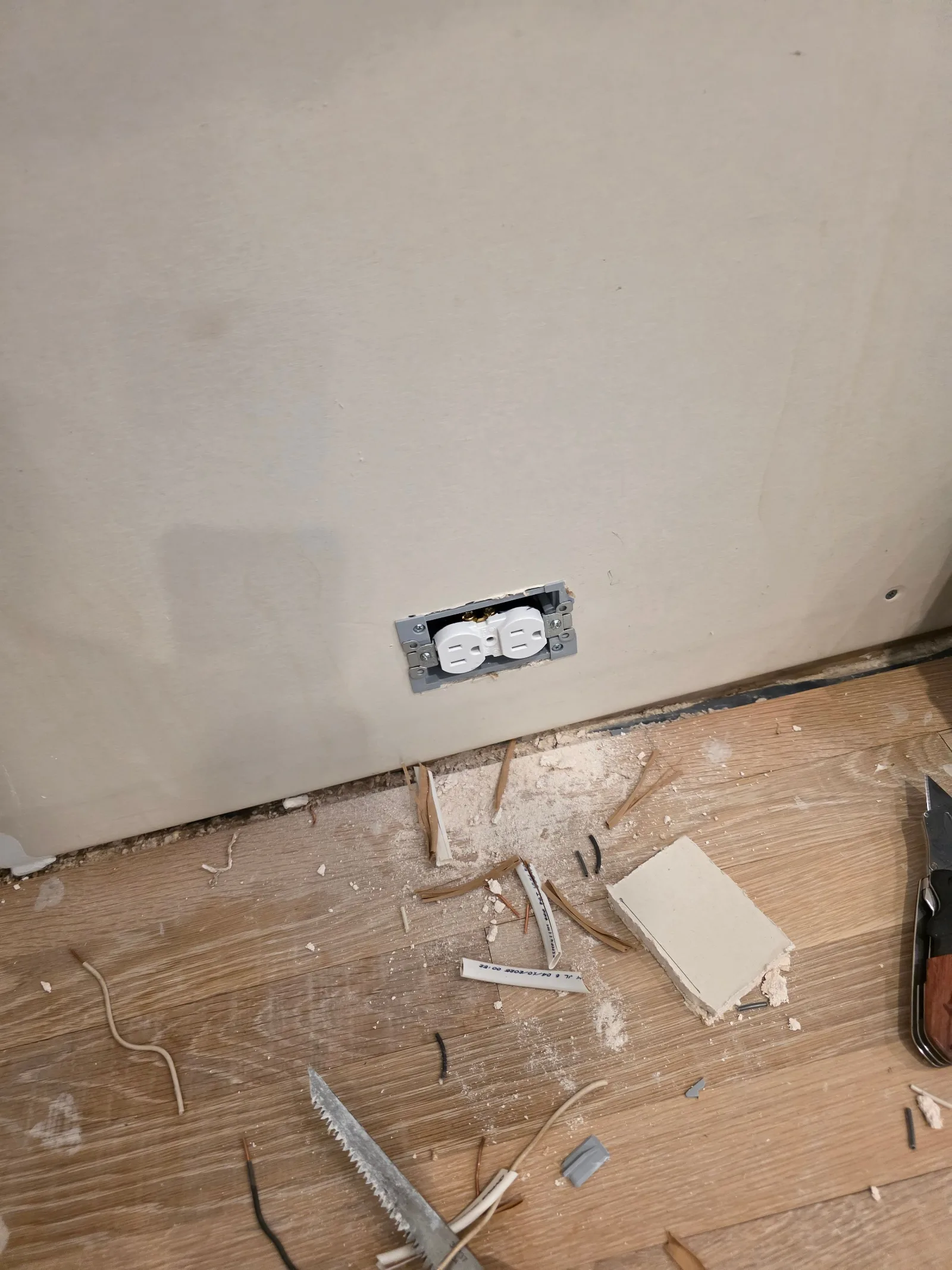 A white duplex electrical outlet installed on a light beige painted wall above a wood floor with debris.