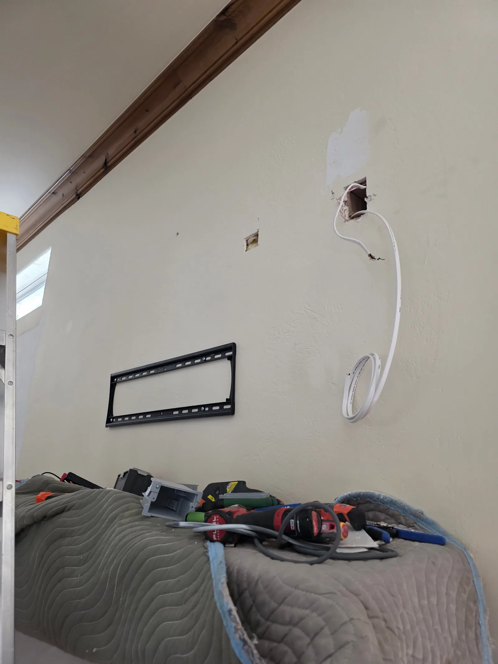 A pale beige wall with a dark wood beam near the ceiling, a mounted TV bracket, exposed wires, and an open electrical box. To