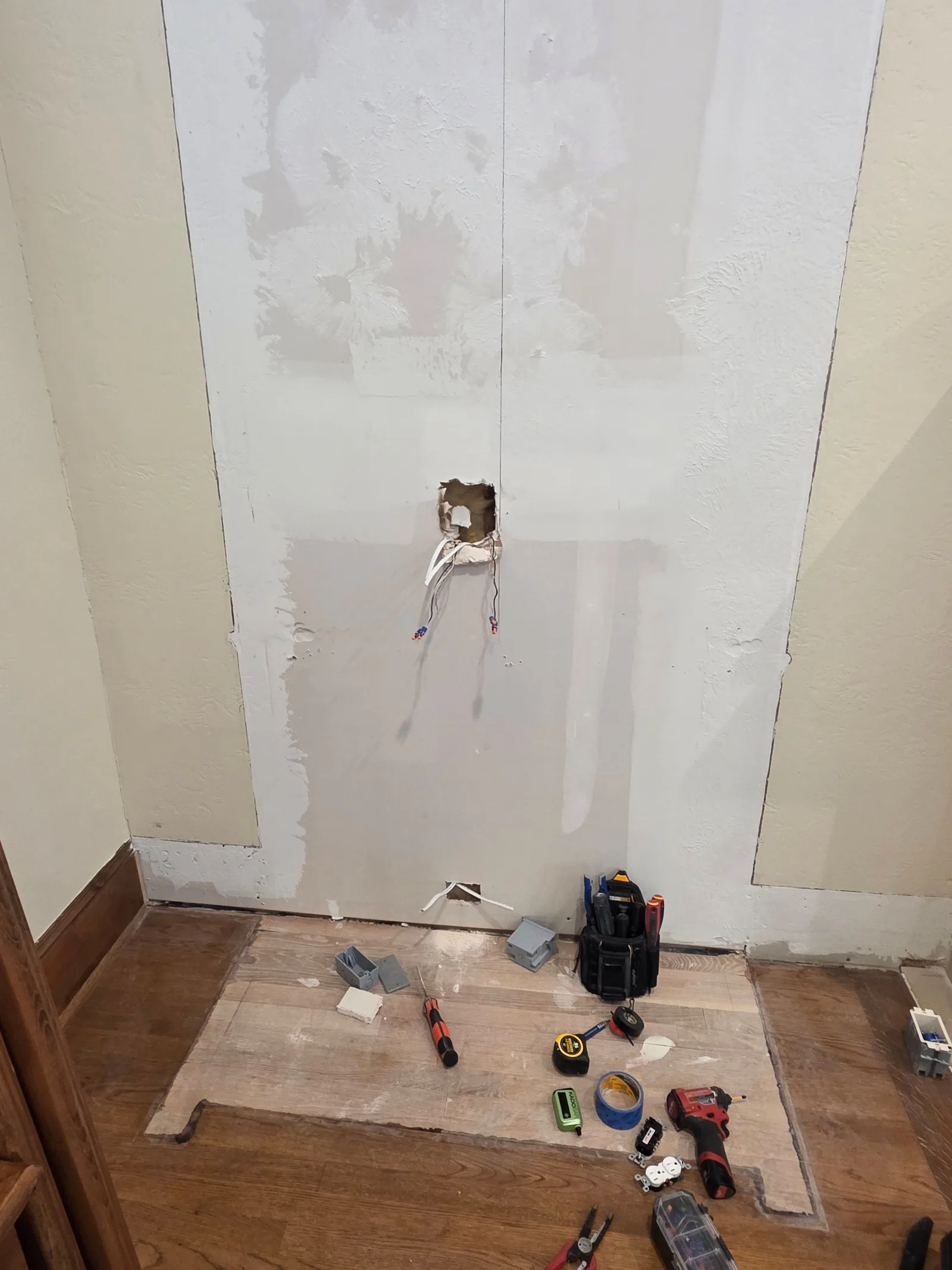 A wall under construction with partially patched drywall, exposed electrical wiring, and wood flooring with a cut-out section