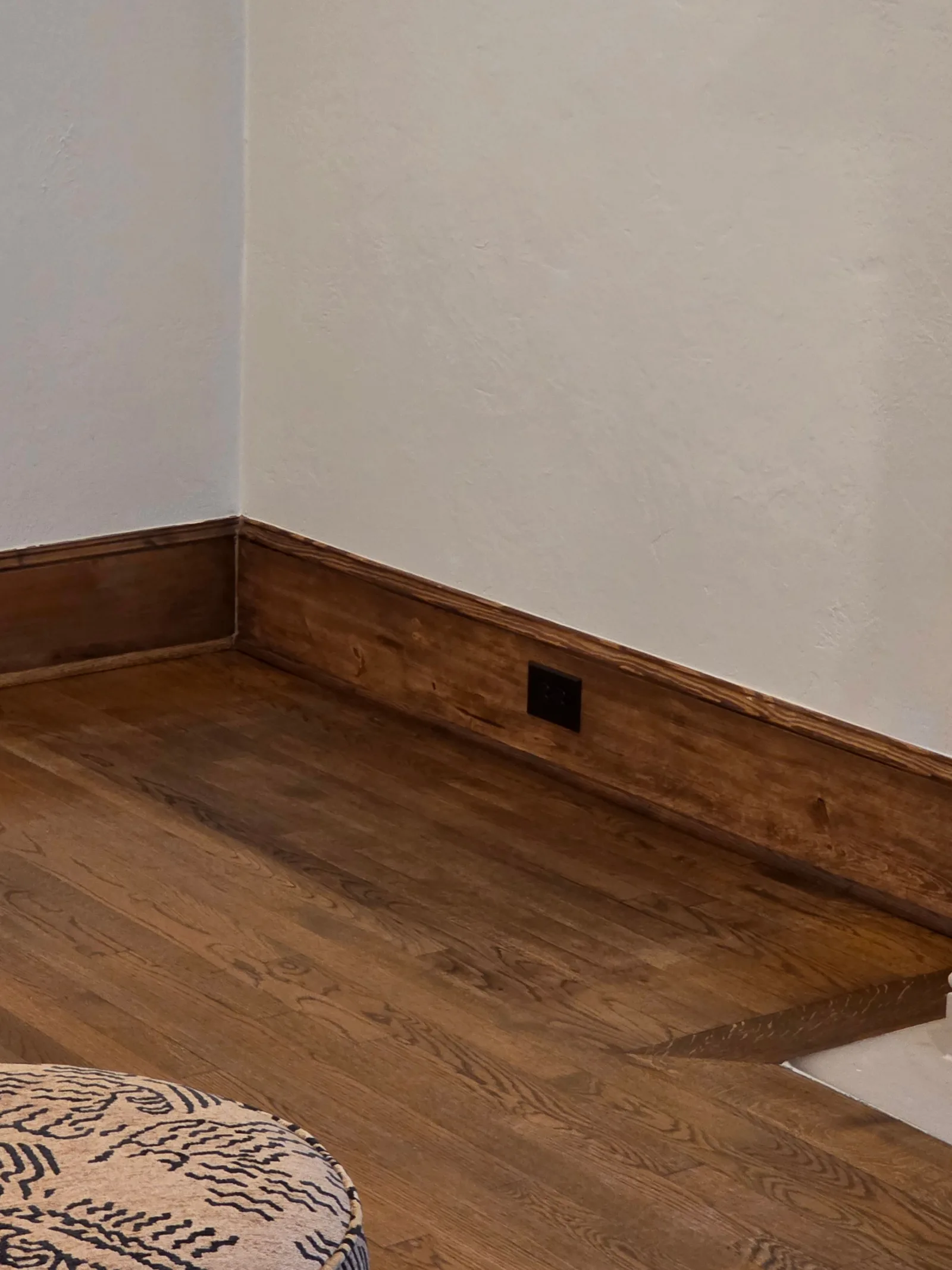 Oak hardwood floor and dark stained wall trim with a recessed electrical outlet, light off-white painted walls.