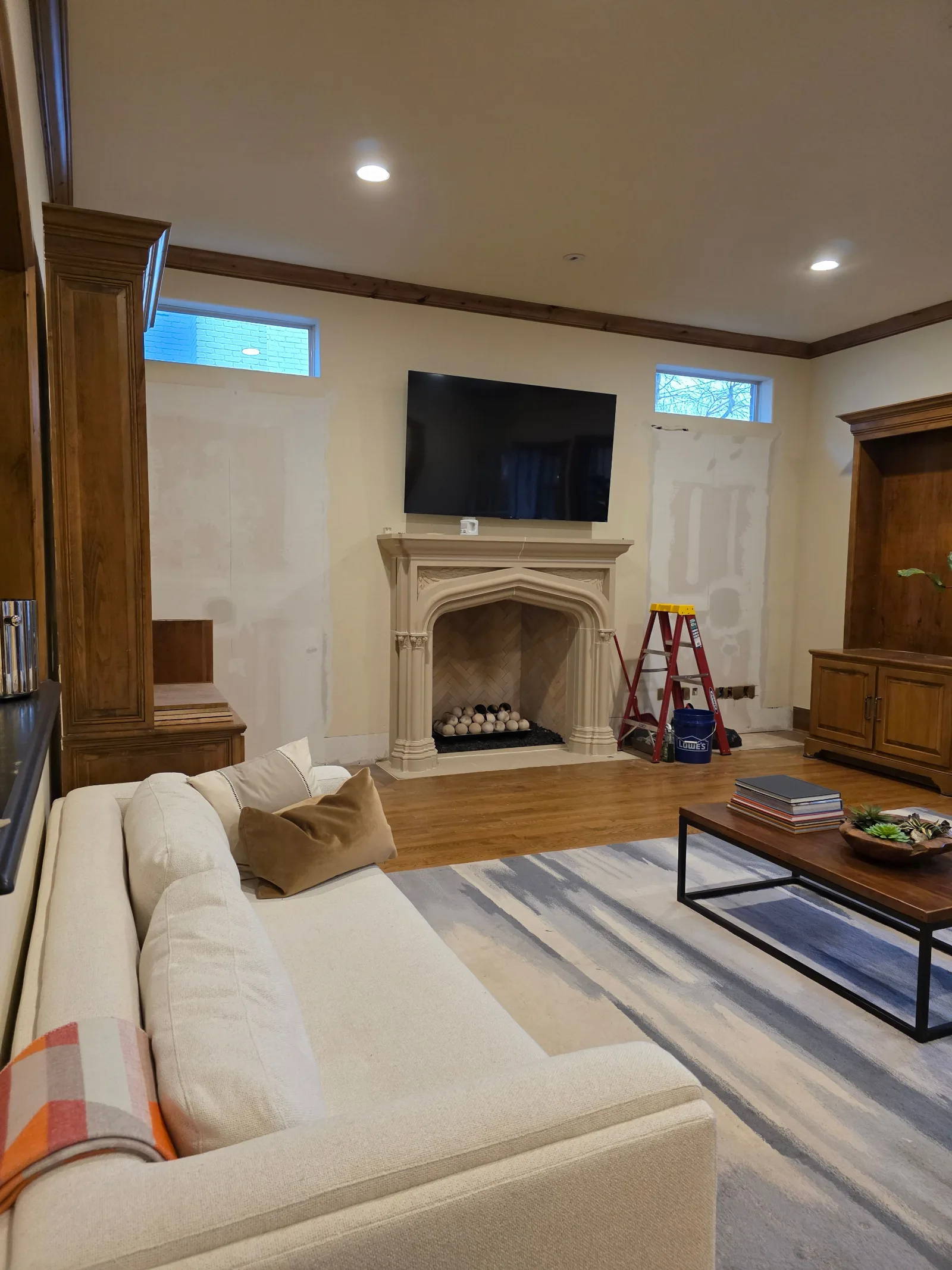 Living room with a large TV above a carved fireplace, hardwood floors, a white sofa, and a painter's ladder. Walls show patch