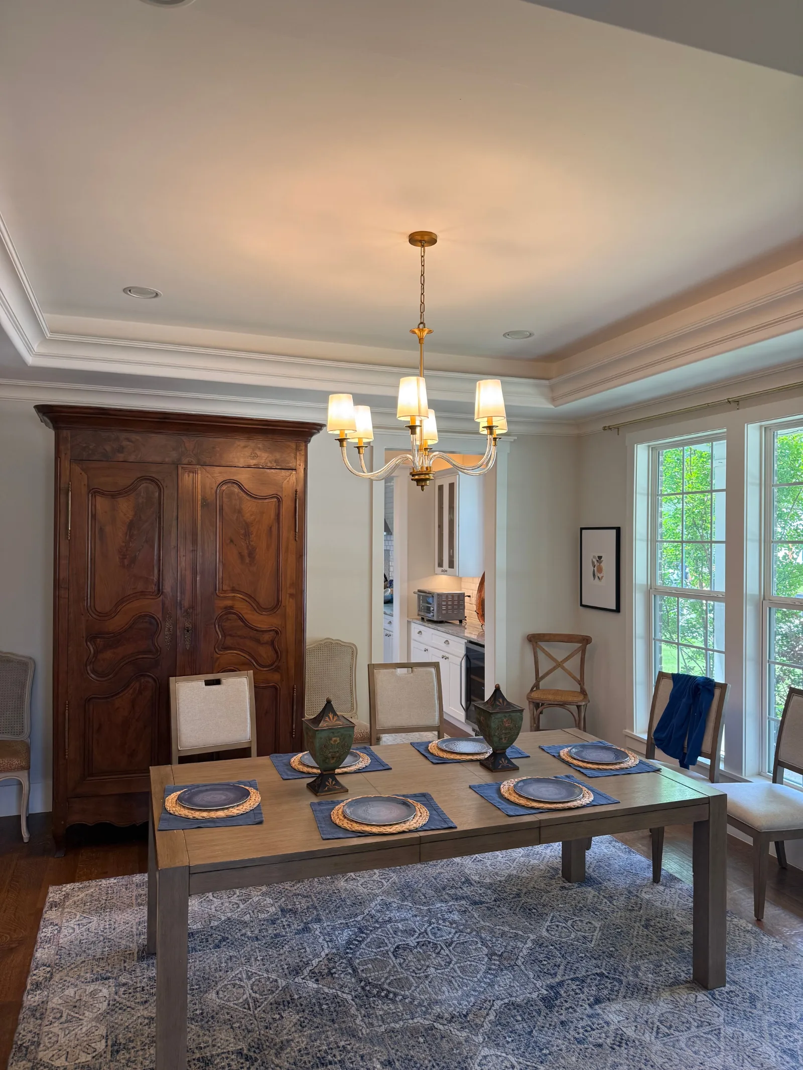 Dining room with a gold and glass chandelier, recessed lighting, and a dark wood armoire. A long dining table with grey chair