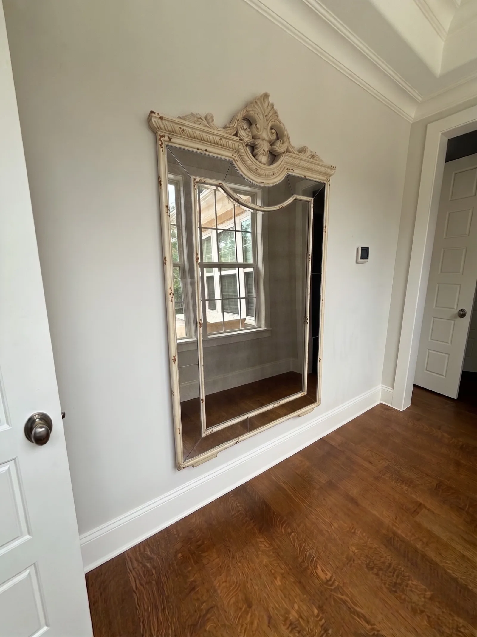 Wall with a large, ornate, distressed antique white mirror reflecting a window. Dark hardwood floors and white trim.