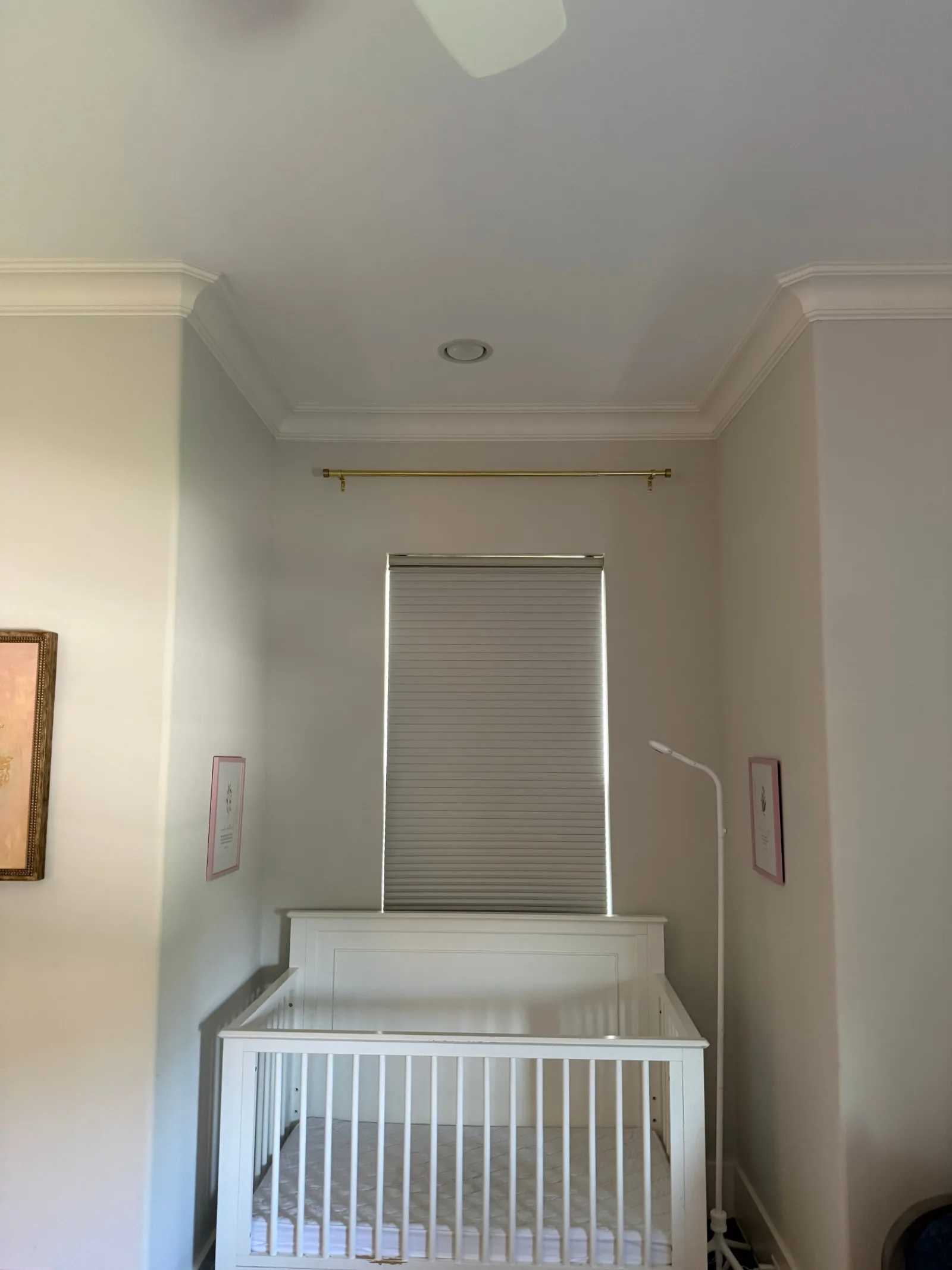 Nursery with an empty white crib below a window with white blinds and a newly installed gold curtain rod. White crown molding