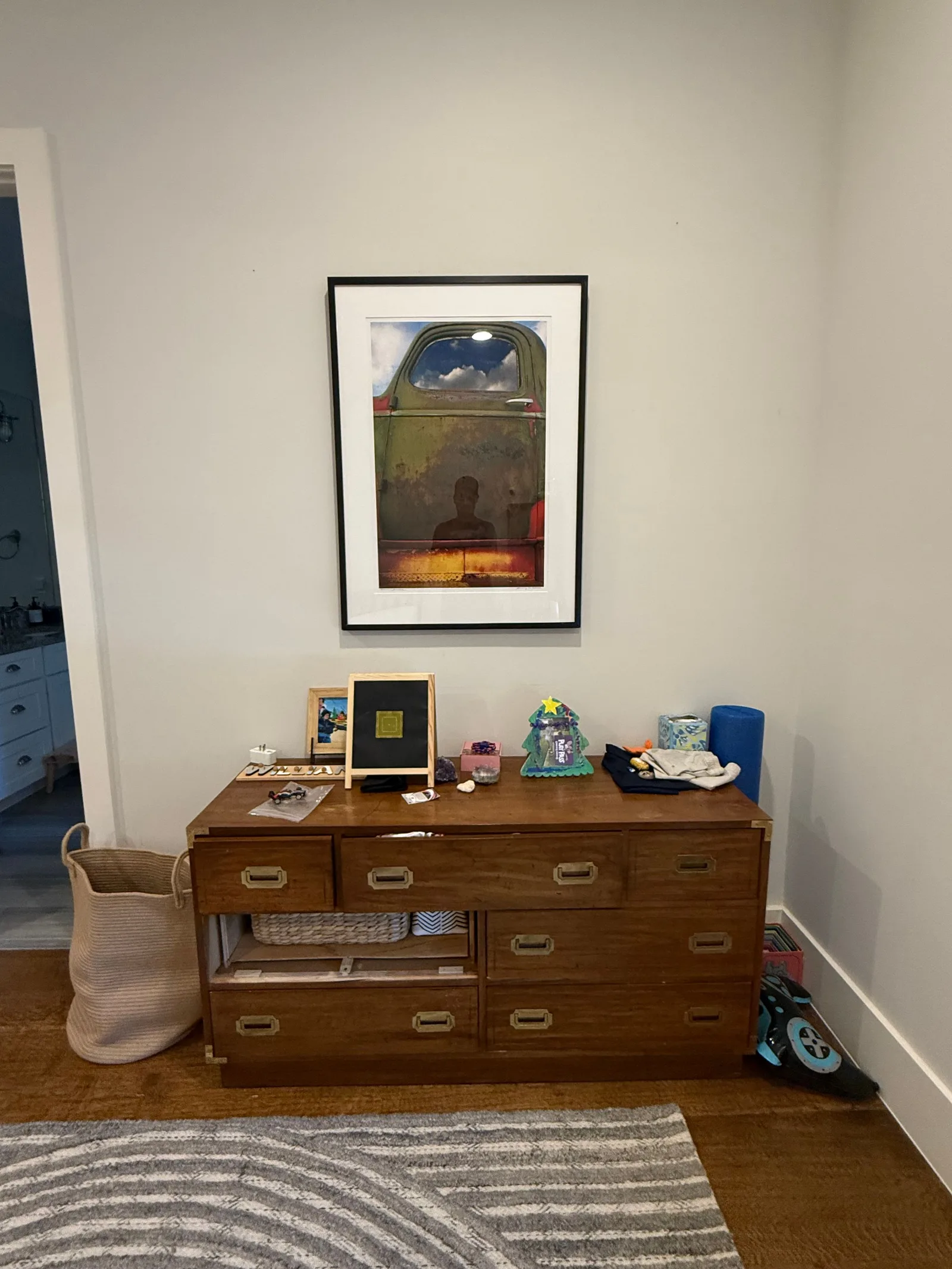 Wood campaign dresser below a framed art print of an old truck, with a tan laundry basket, yoga mat, and rug on hardwood floo