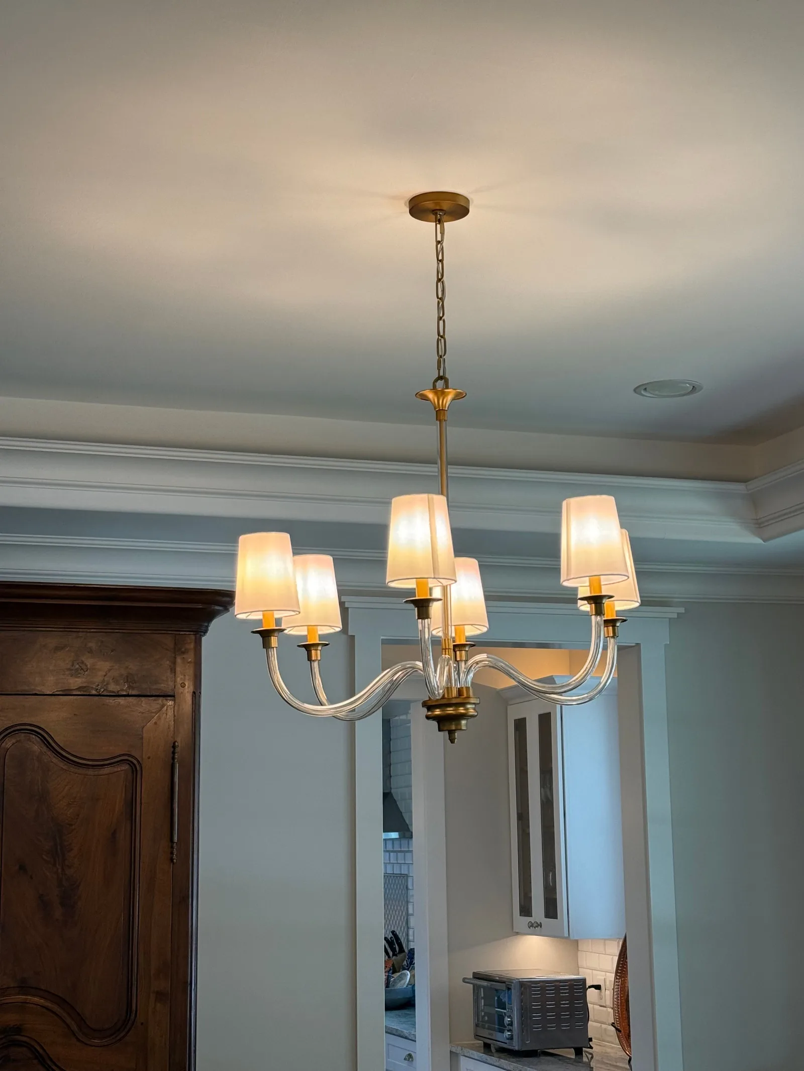 A modern chandelier with 6 white-shaded lights and a brass base hangs from a white ceiling with crown molding. A dark wood ca