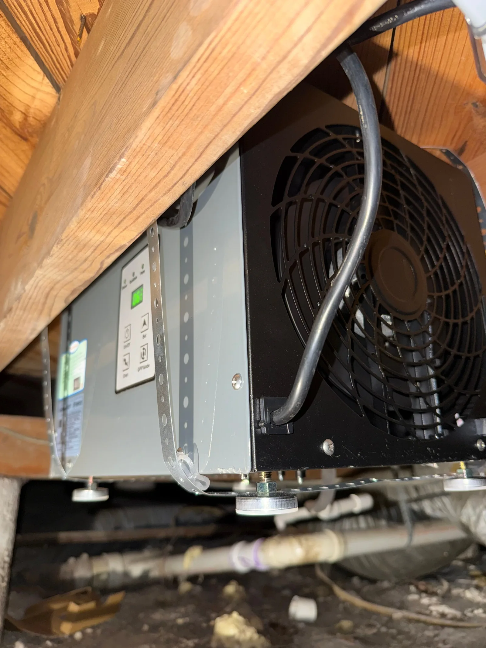 Dehumidifier with a gray body, black fan casing, and control panel, mounted under a wooden floor joist in a crawlspace.