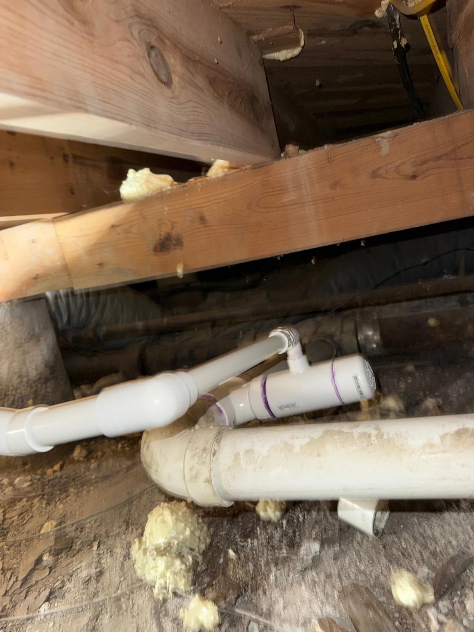 Under-home view of crawlspace structural beams, insulated white plastic pipes, yellow and black electrical wiring, and spray