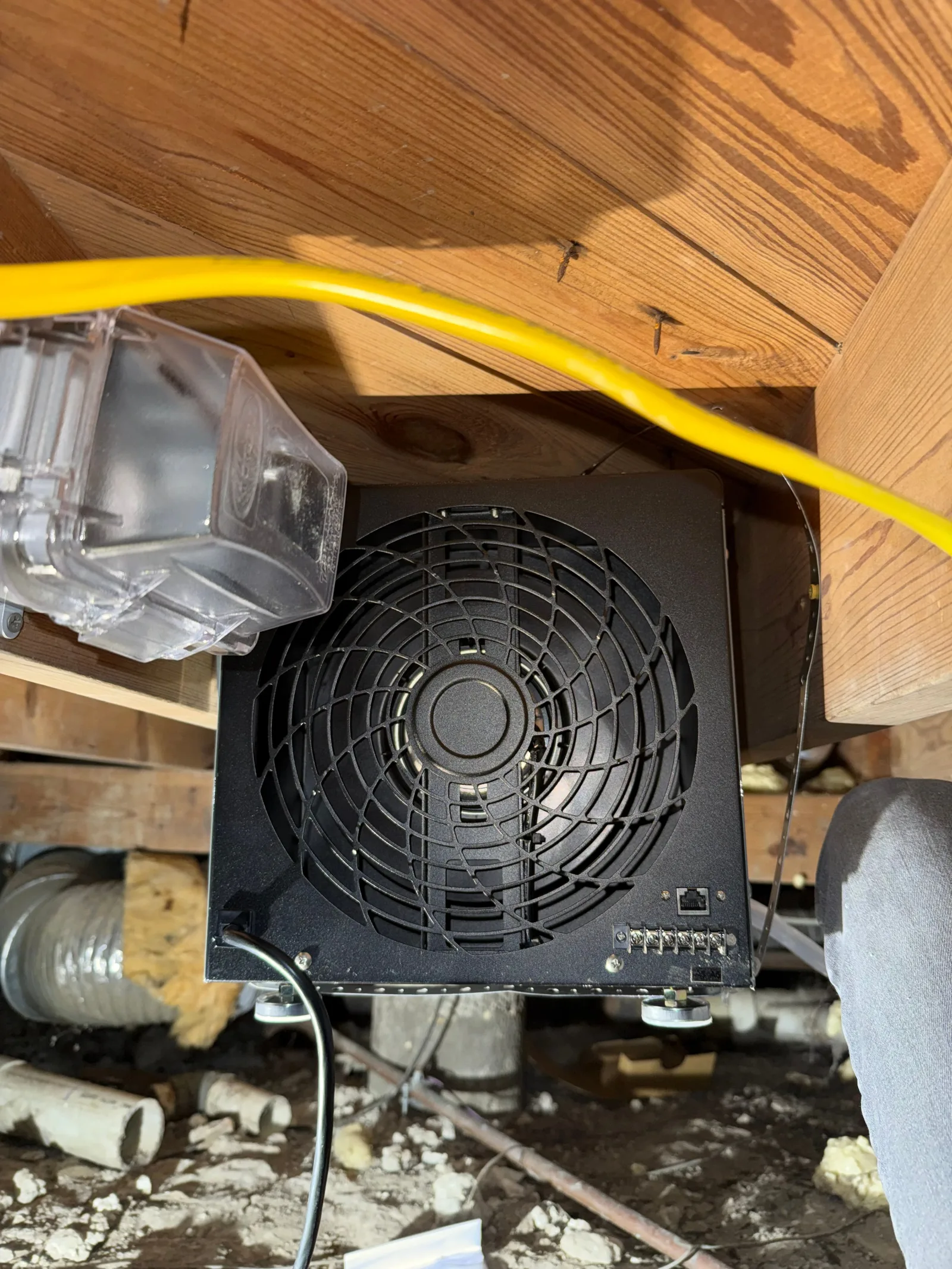 A black crawlspace dehumidifier is securely mounted to wooden beams, with visible wiring and a clear plastic cover.