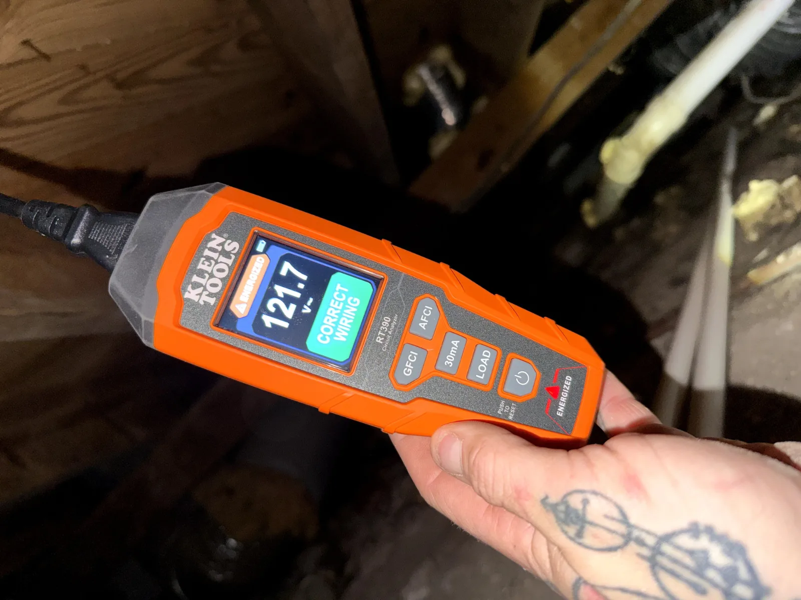A hand holding an orange Klein Tools circuit analyzer in a dark crawlspace, showing "121.7 V~" and "CORRECT WIRING".