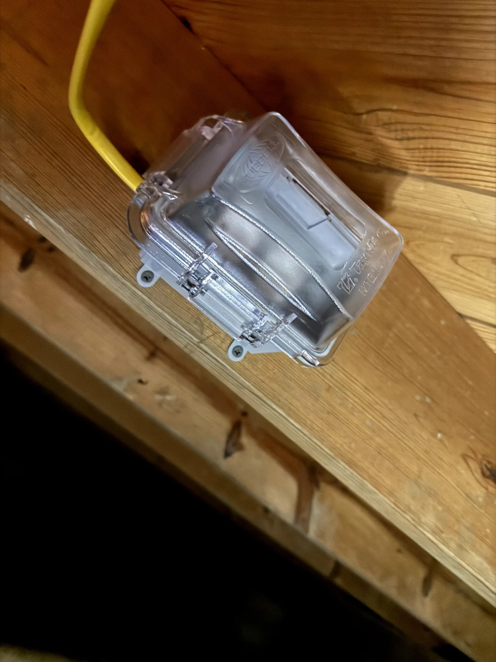 Close-up of a clear plastic outlet cover box with yellow electrical conduit attached, screwed to a light brown wooden joist.