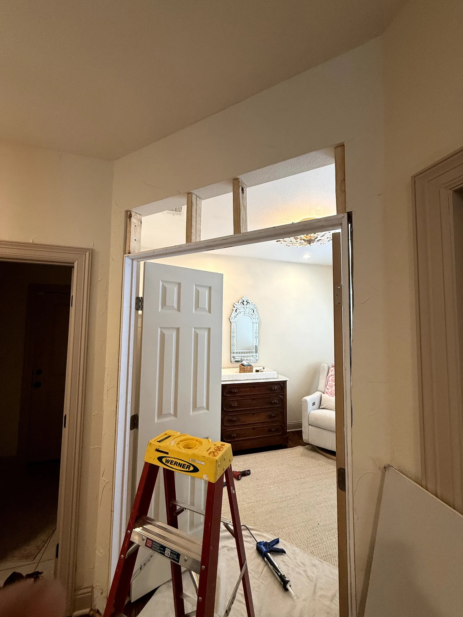 Entryway to a renovated room with a white door partially open. A wooden dresser, mirror, and white armchair are visible insid