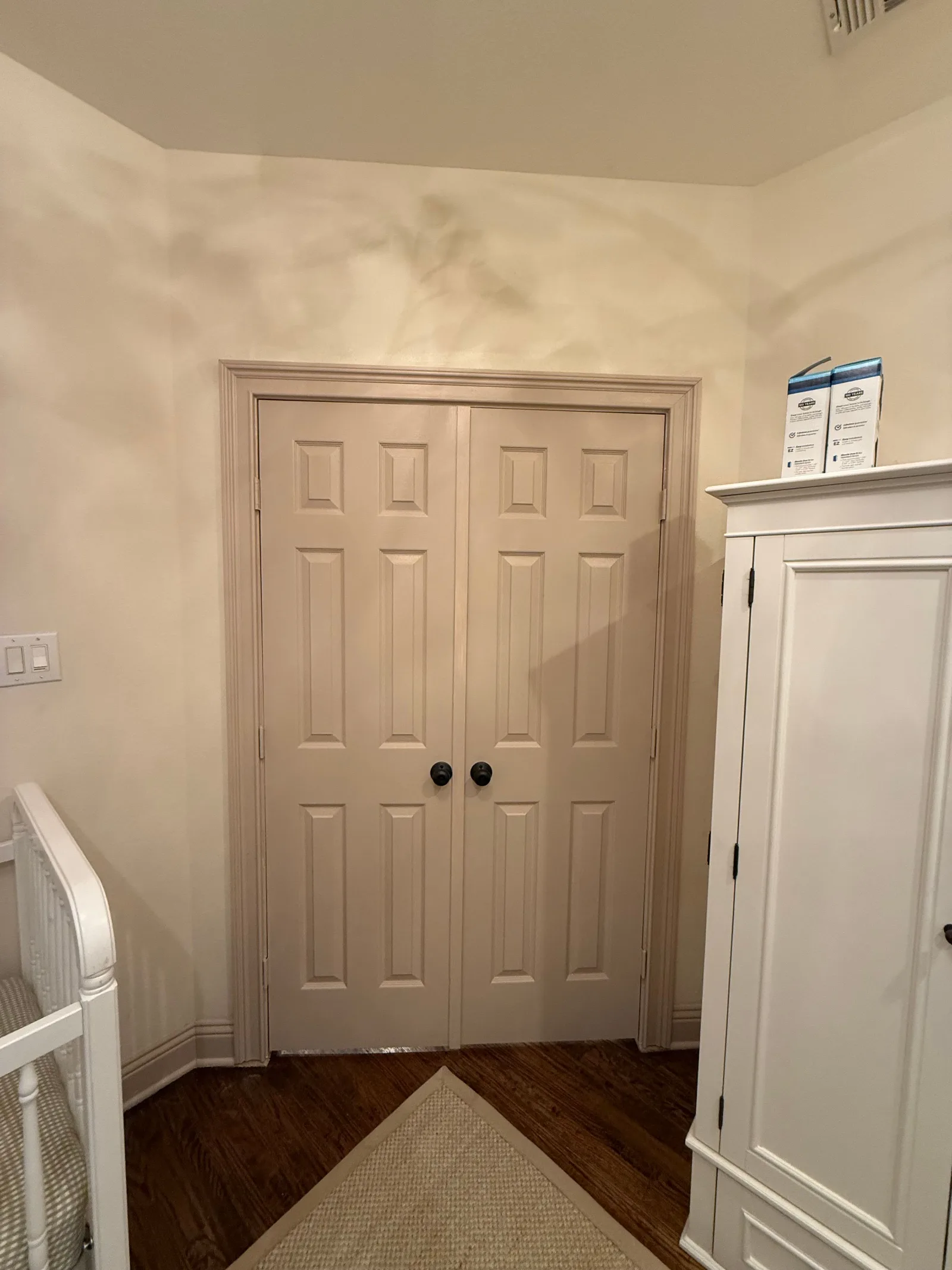 Nursery with cream walls showcasing newly installed French doors with dark round knobs, wood floors, and a white crib.