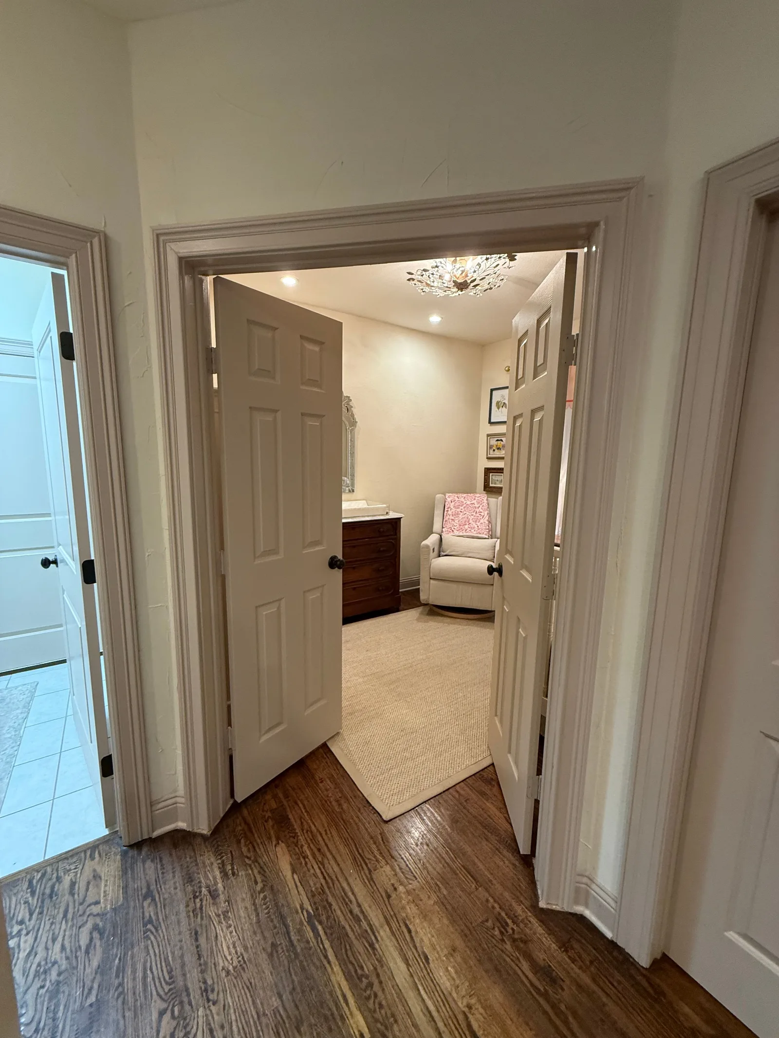 Double French doors open into a nursery with a beige rug, white rocking chair, brown dresser, and decorative chandelier.