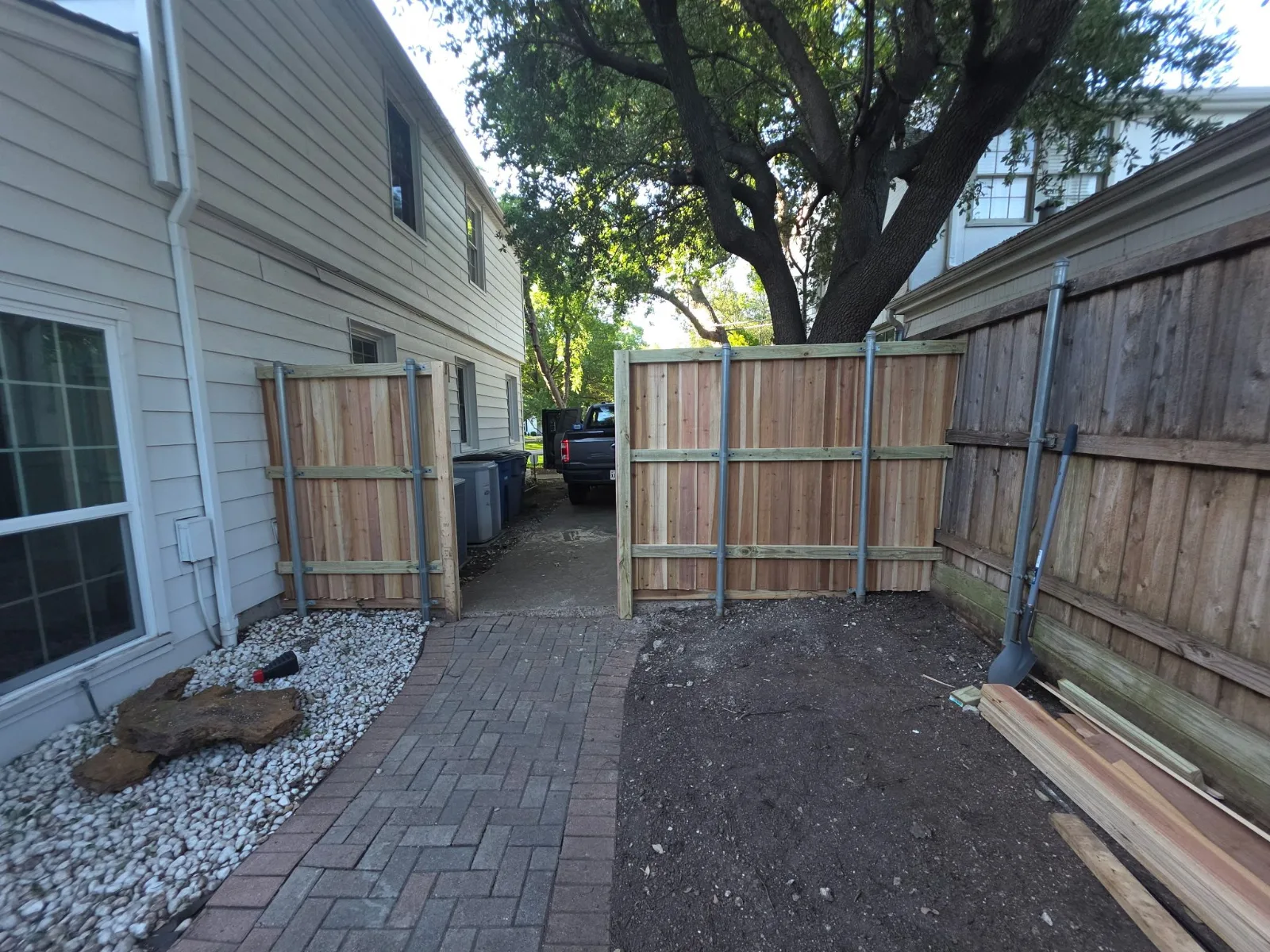 Backyard view showing a newly installed wooden fence, a white siding house, a paved brick pathway, and a gravel area.