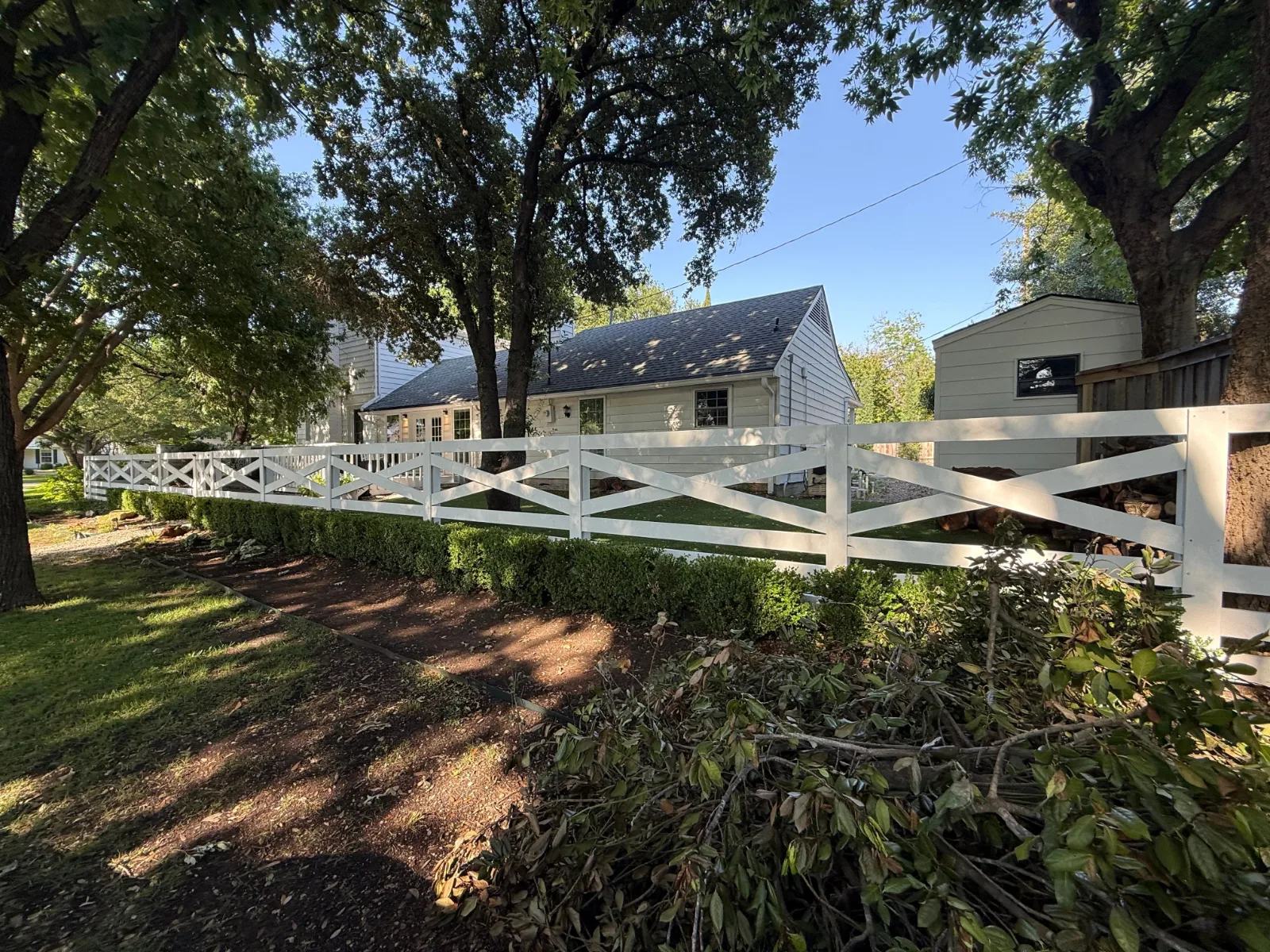 A white house with a dark roof and white trim behind a long, white criss-cross fence, lined with green bushes.
