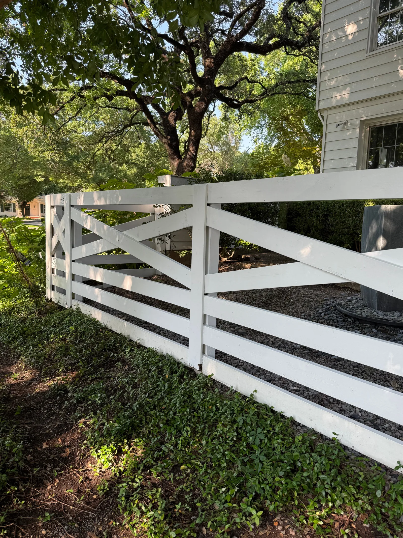 A newly installed white wooden fence with a gate, surrounded by green ground cover and mature trees, next to a white house.