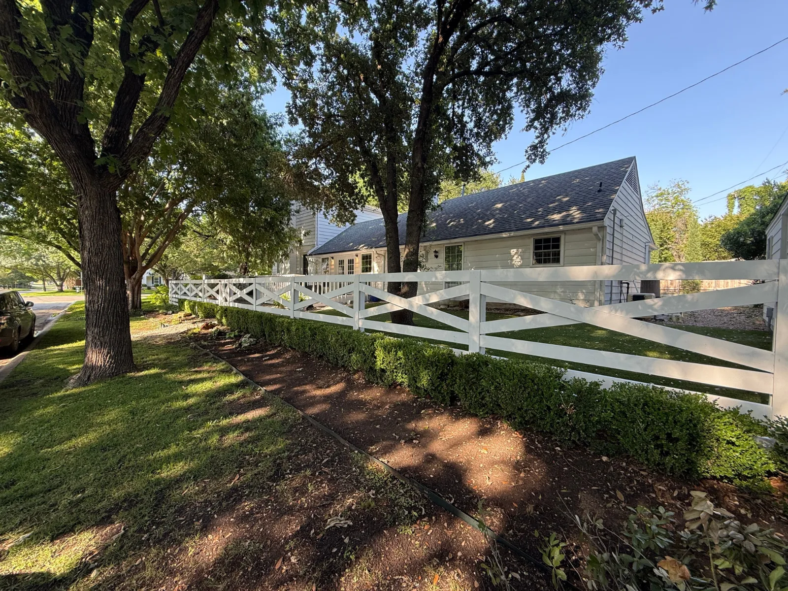 White picket fence with diagonal crosses, evergreen shrubs, and a white house with dark roof and windows in the background.