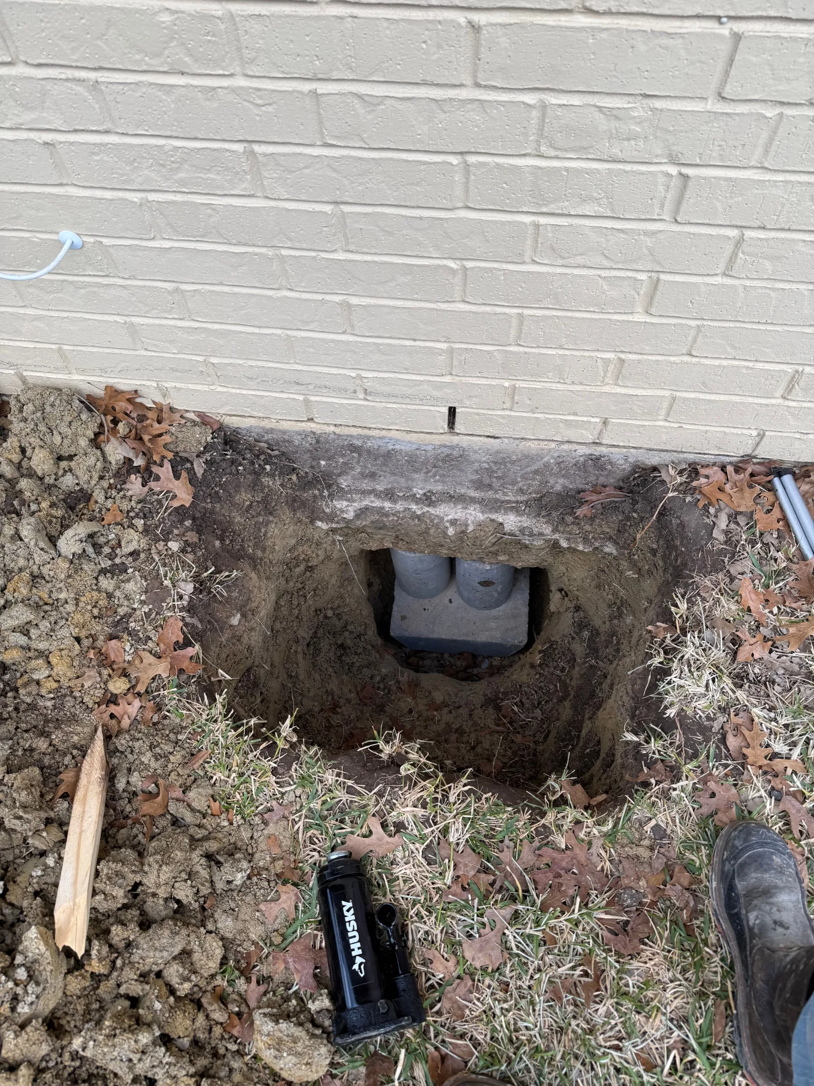 Foundation repair work: a deep pit next to a light-colored brick house, showing concrete piers installed beneath the foundati