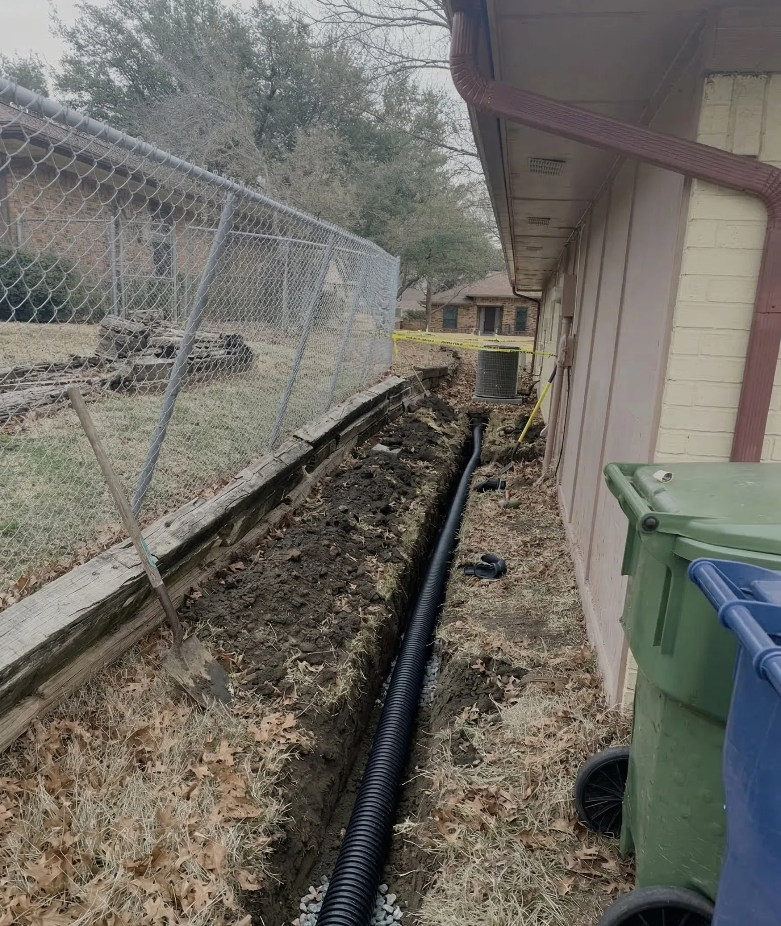 A black corrugated drainpipe laid in a narrow, muddy trench alongside a house foundation with exposed white brick.