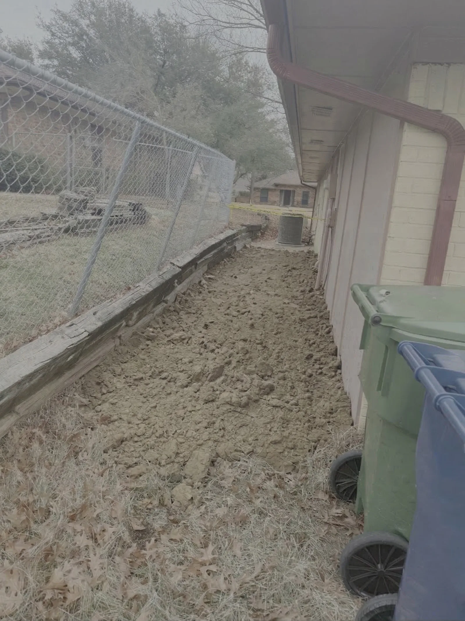 Side view of a house with light yellow siding and a brown gutter, showing freshly excavated dirt beside the foundation.