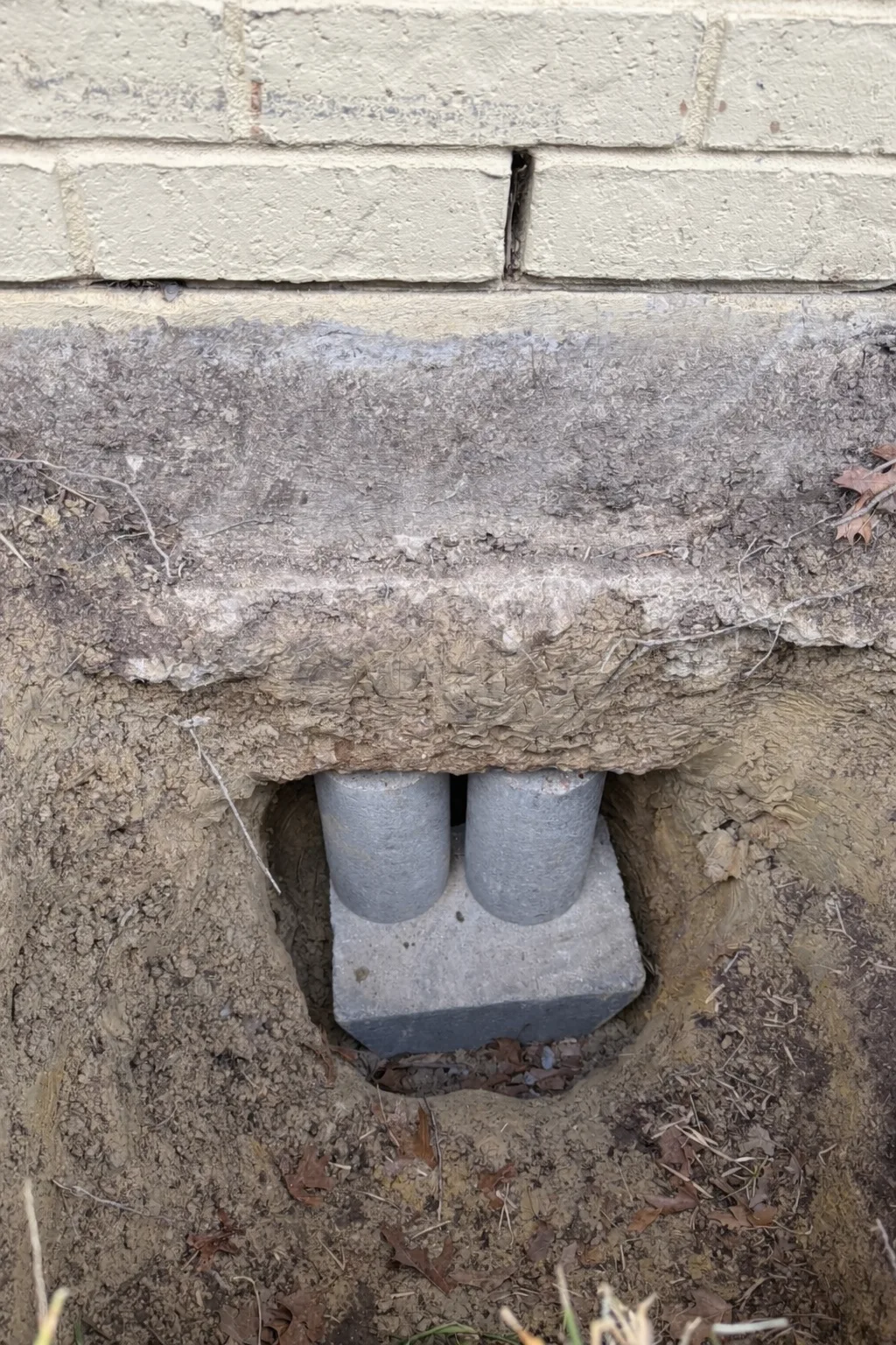 Two cylindrical concrete piers on a rectangular base in a dug pit, supporting a foundation with cracked, painted brick.