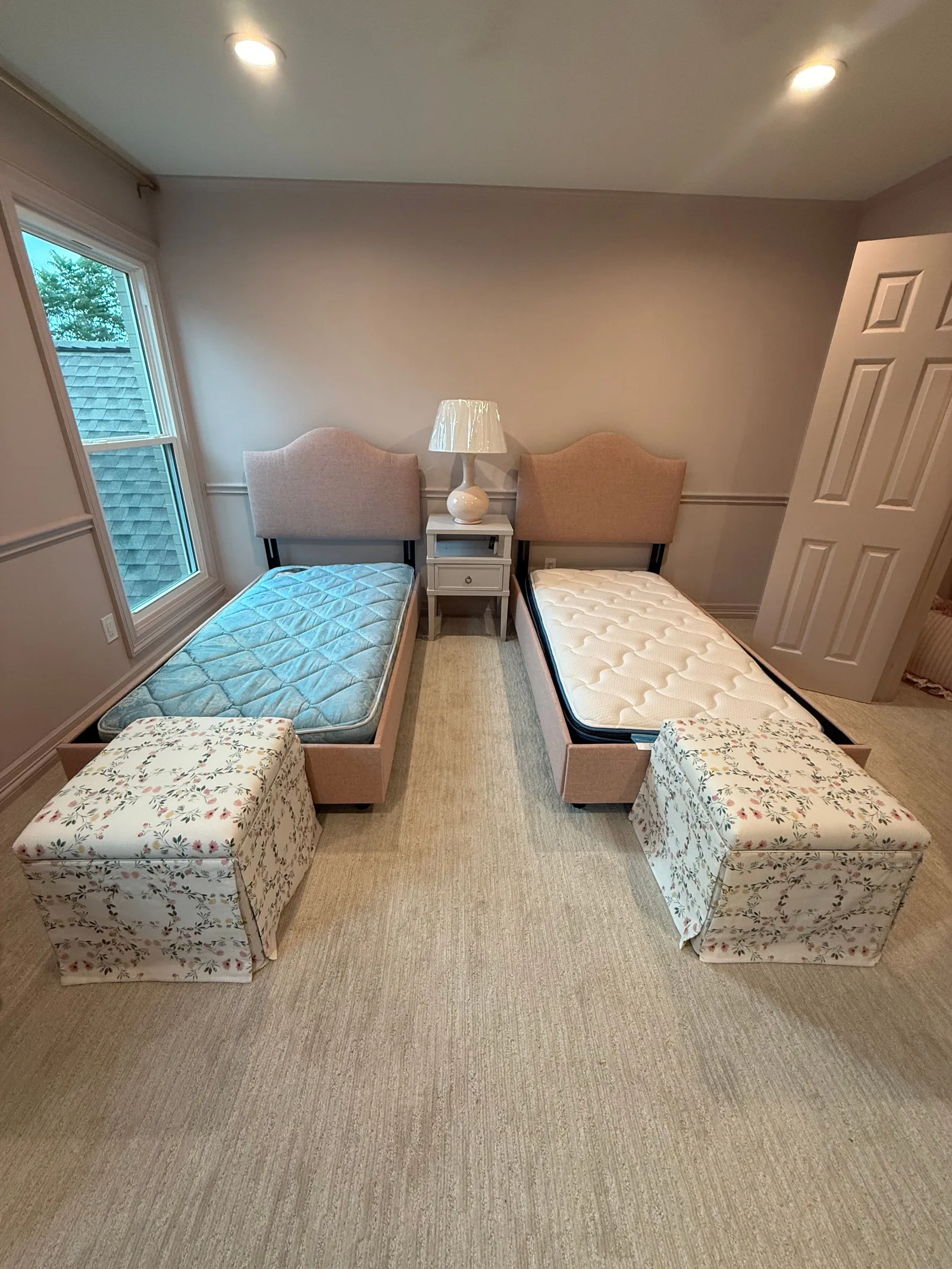 Two pink upholstered twin beds with mattresses, a white nightstand, and two floral footstools in a bedroom with light pink wa