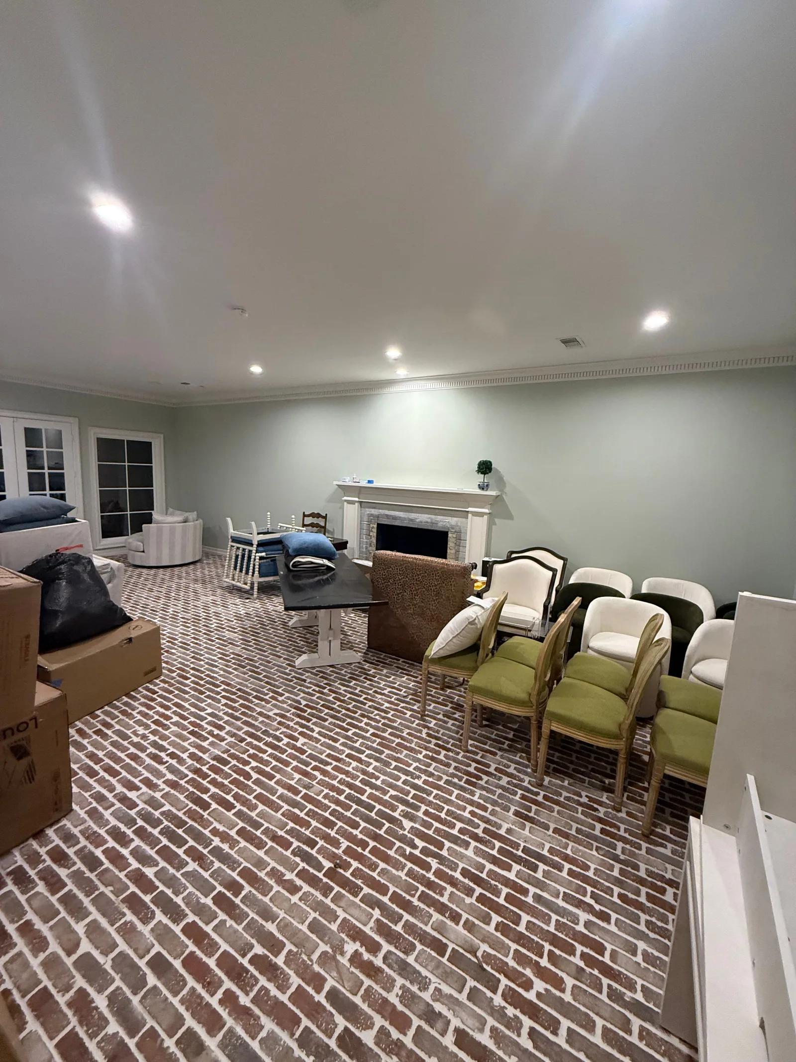 A spacious living room with brick flooring and light green walls, cluttered with various unassembled furniture, boxes, and a