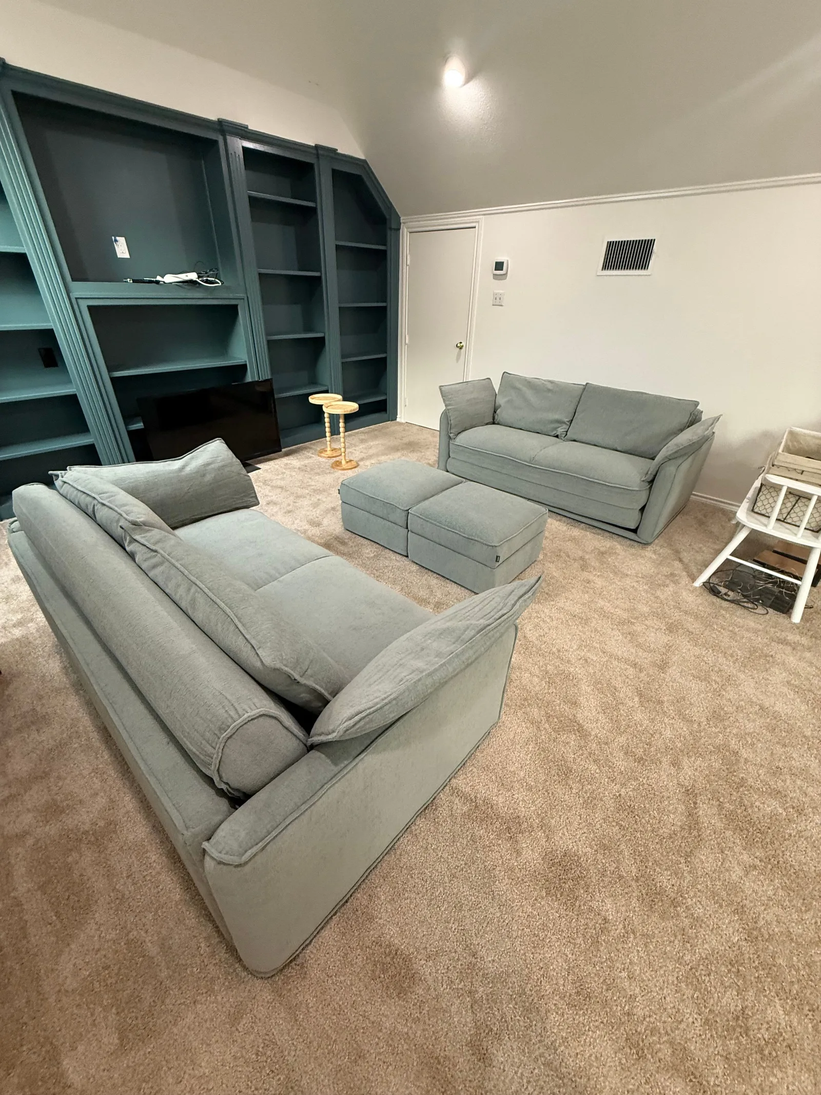 Two light gray sofas and two ottomans on a beige carpet, facing dark blue built-in bookshelves with a TV.