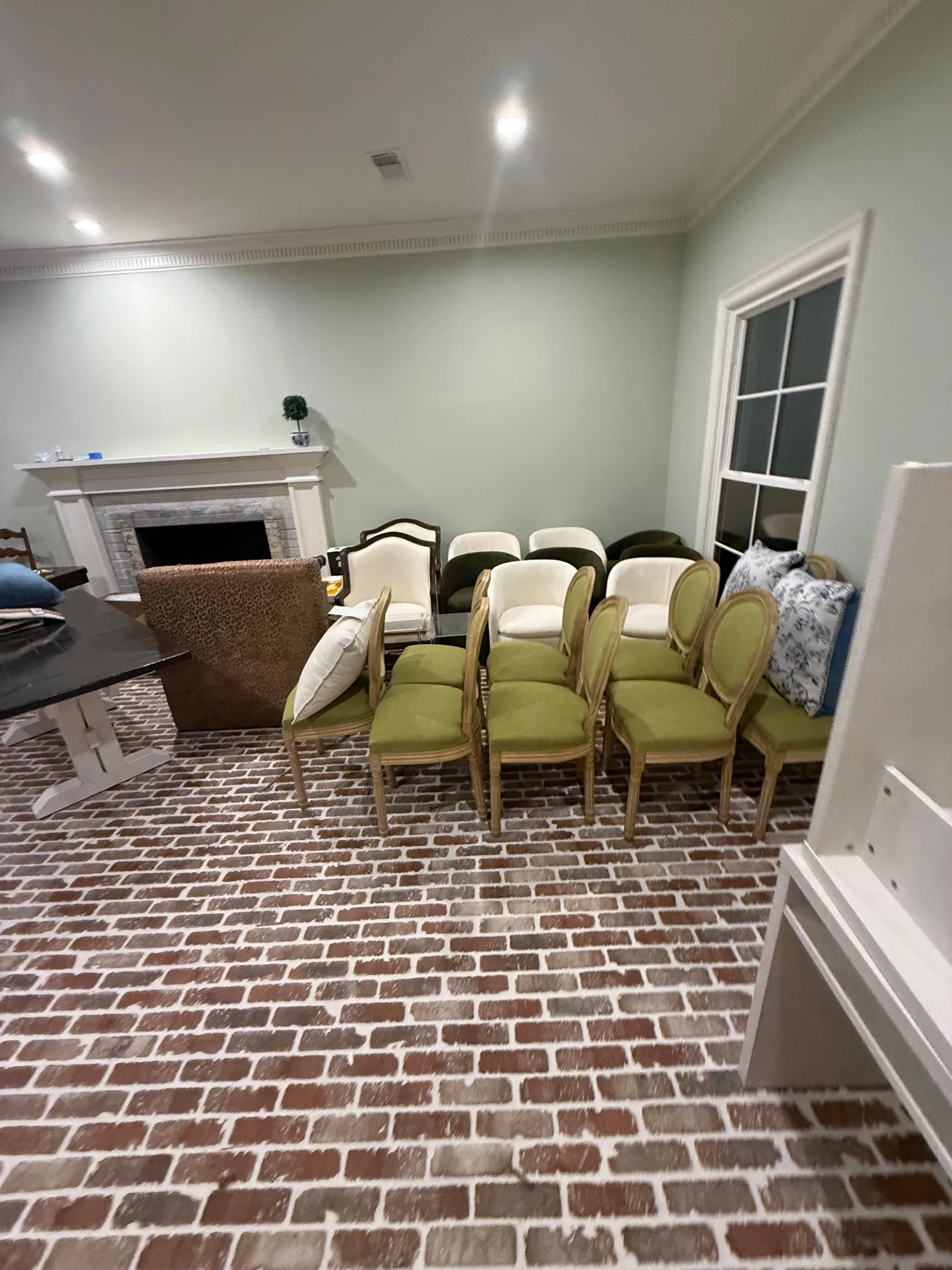 Living room with light green walls and reclaimed brick flooring. A white fireplace, dining table, and various chairs are pres