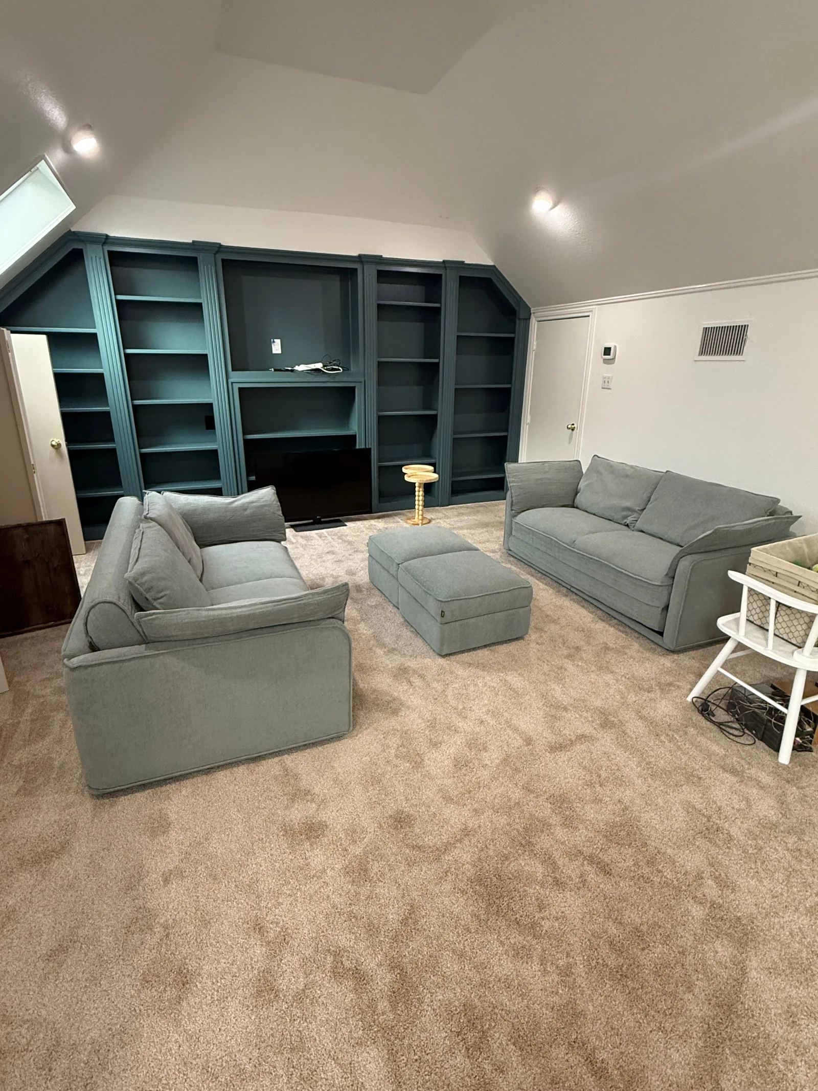 A living room with two light grey sofas, a matching ottoman, and a large custom teal built-in shelving unit.