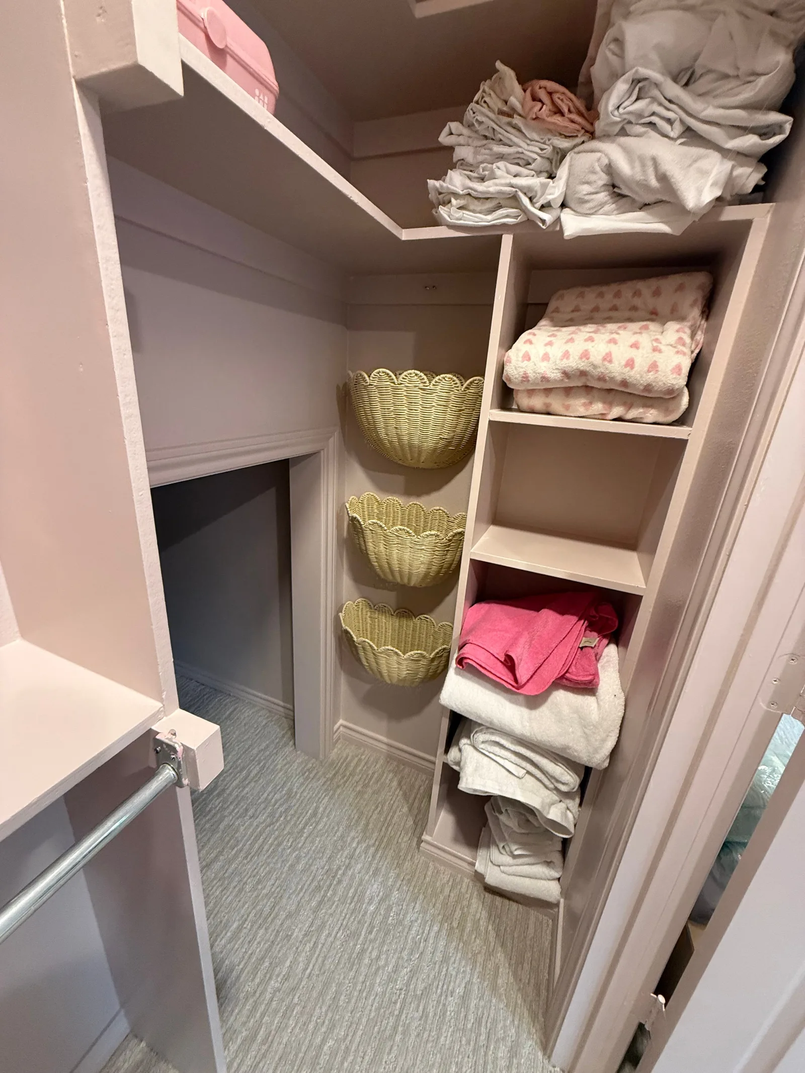 A pale pink closet interior with built-in shelving, a clothes rod, three woven wall baskets, and plush towels.