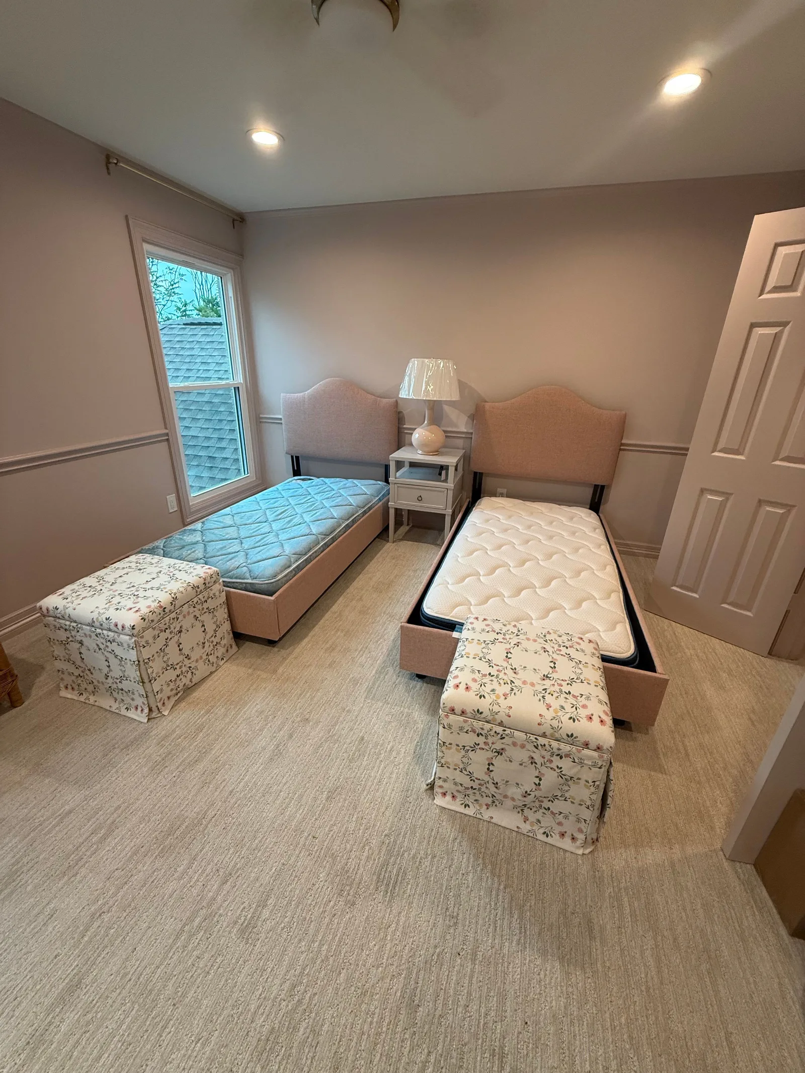 Two pink upholstered twin beds with headboards and mattresses in a light-colored bedroom with neutral carpet and an unclothed