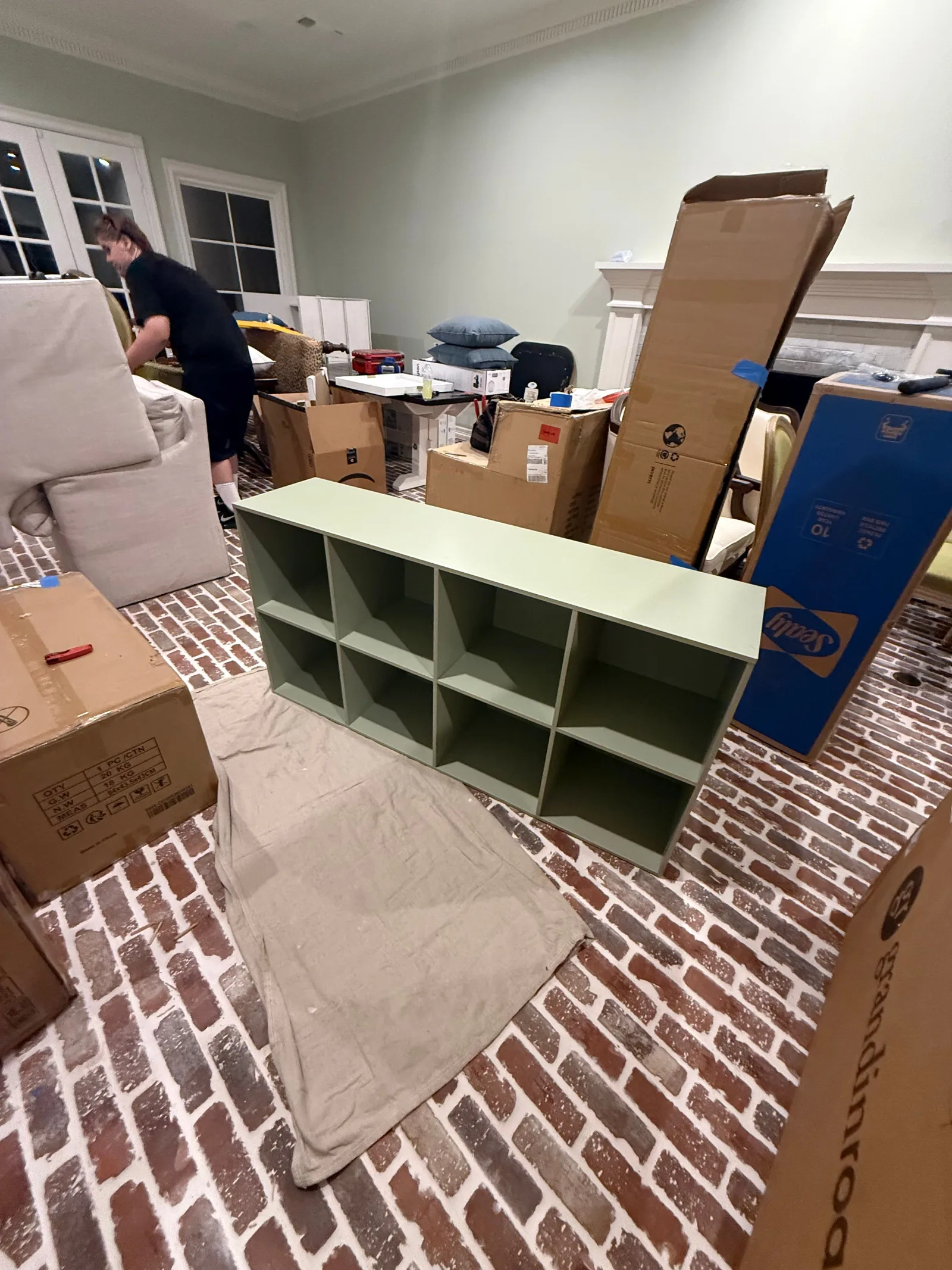 A pale green modular shelf unit on a drop cloth, surrounded by boxes and unassembled furniture in a room with brick flooring.