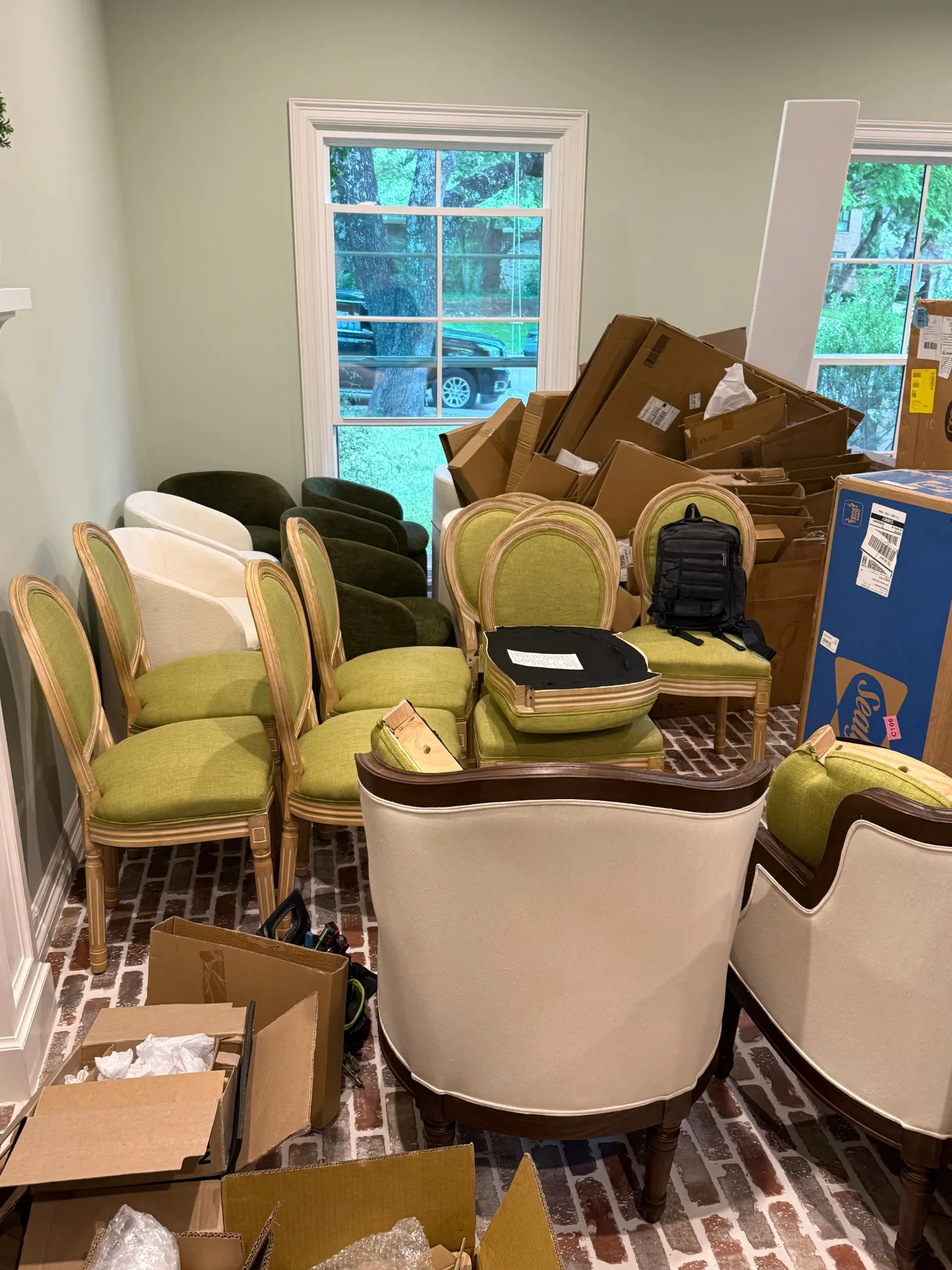 A chaotic room with a green-painted wall, brick flooring, and multiple green chairs, white chairs, and brown cardboard boxes.