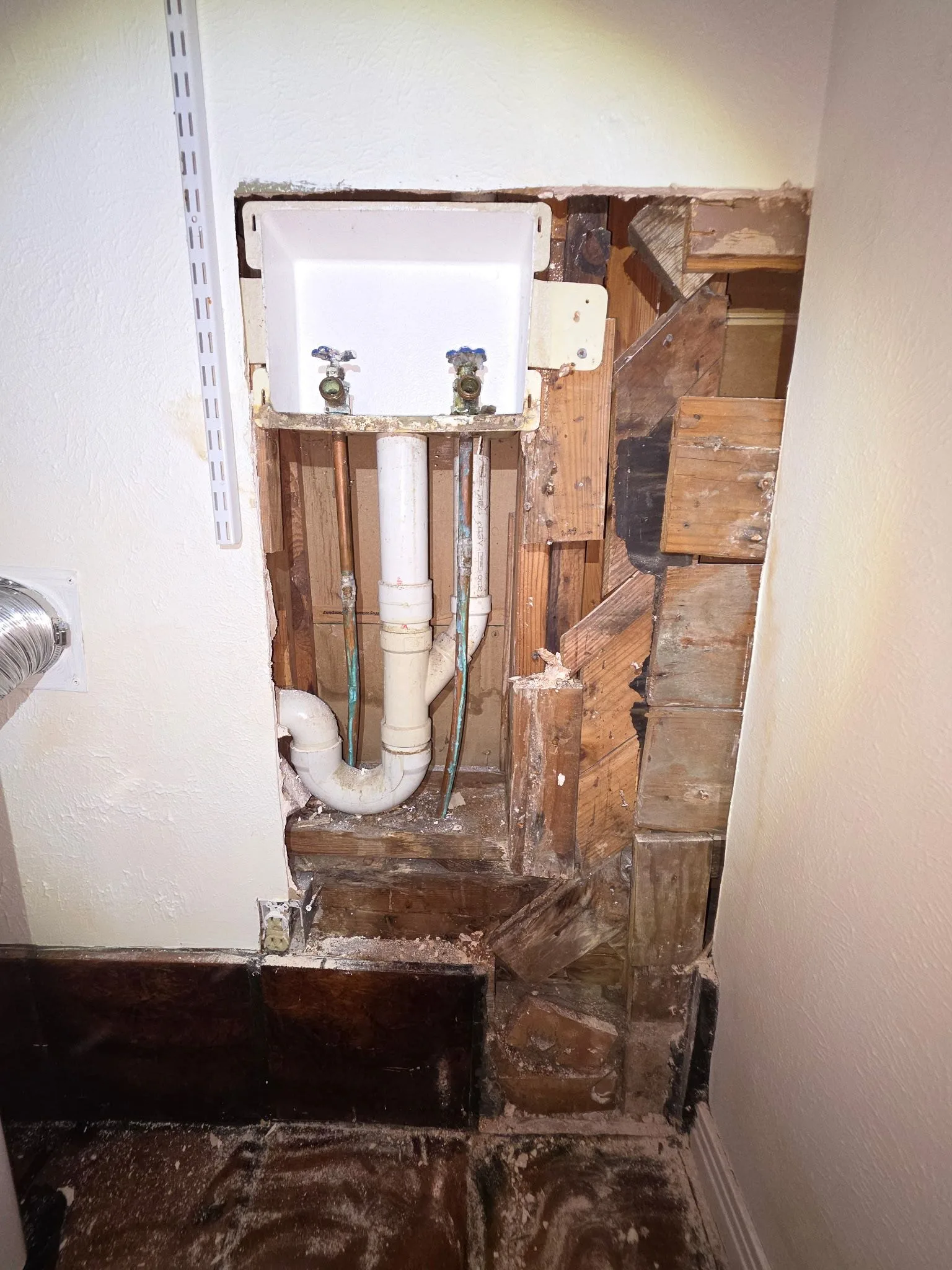 Damage to a laundry room wall exposing wooden studs, white plumbing pipes, and copper pipes for a washer connection.