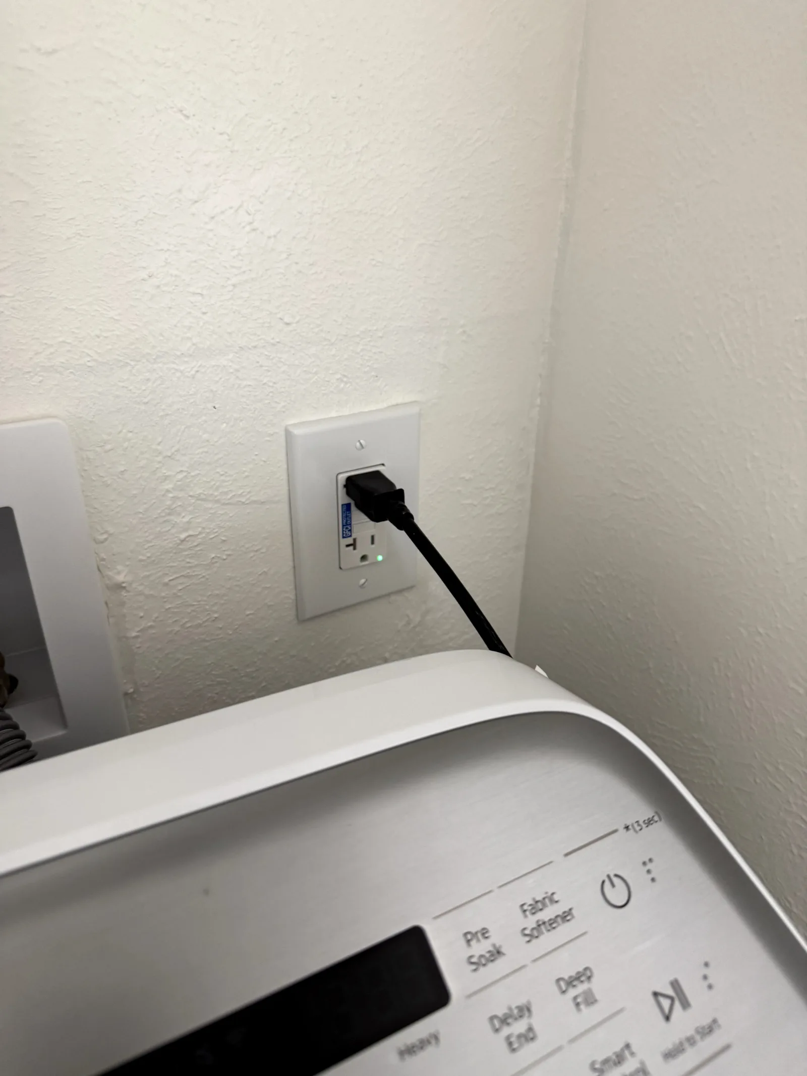 Top of a washing machine with visible controls, a wall outlet, and a black power cord plugged in.