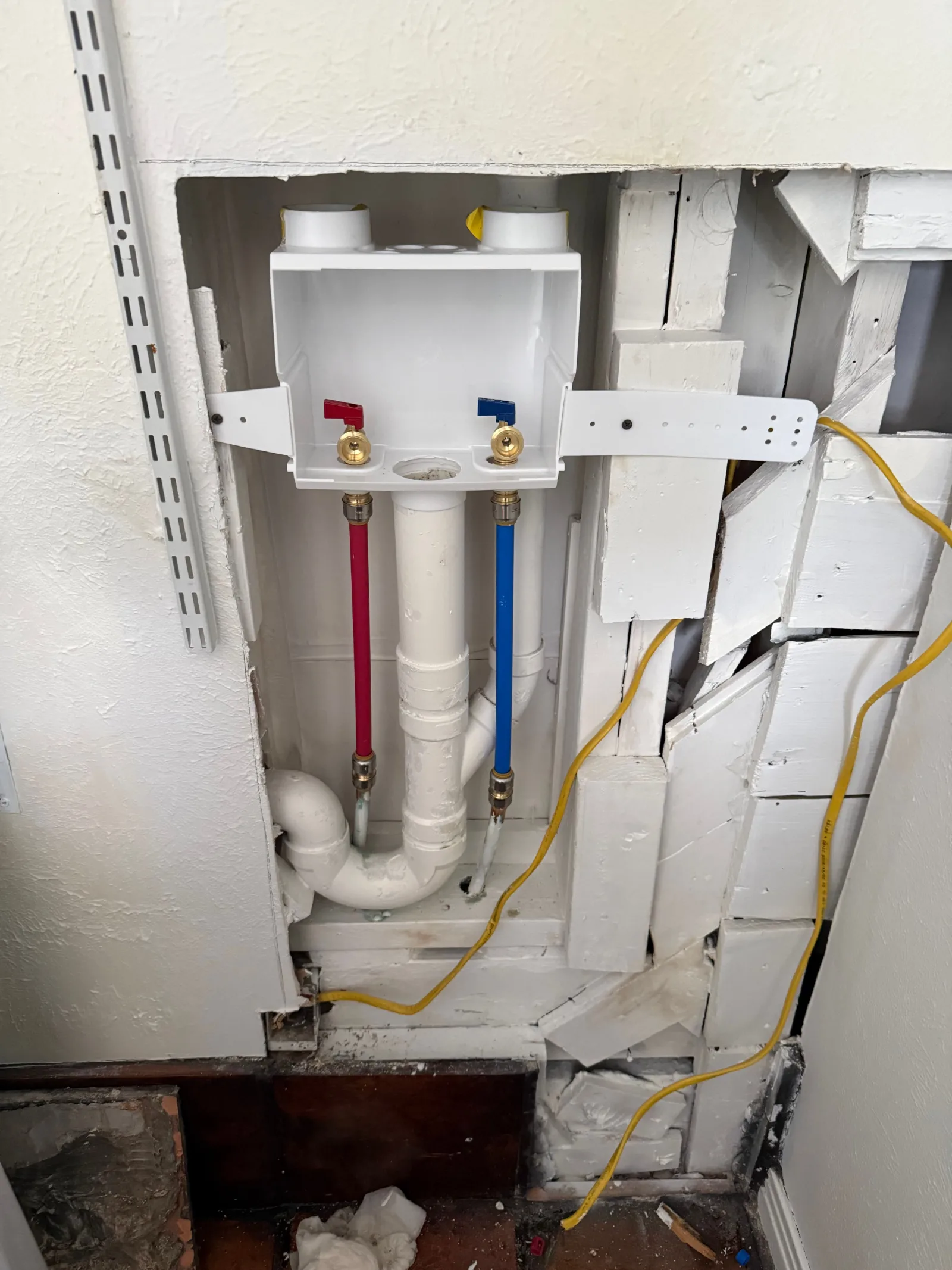 A laundry room's unfinished wall reveals water hookups, drain pipes, electrical wiring, and exposed wood studs.