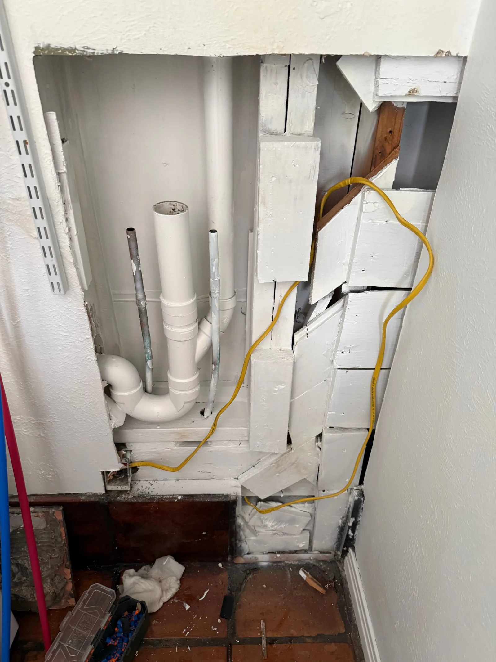 Damaged laundry room wall with exposed white PVC pipes, metal plumbing, wood lath, and a yellow electrical wire.