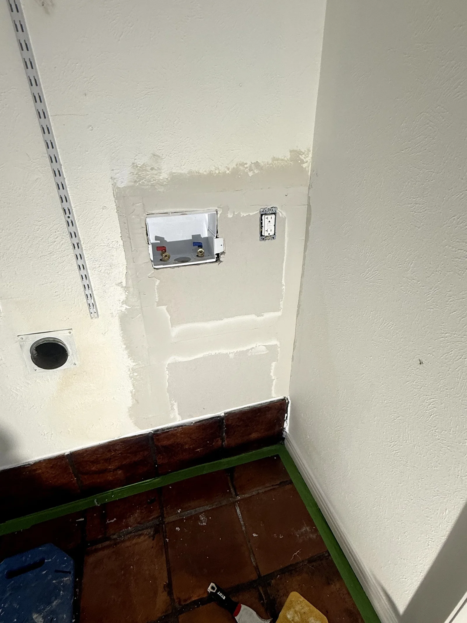 A laundry room with unfinished white walls, a laundry box, an outlet, and a vent hole above terracotta floor tiles.