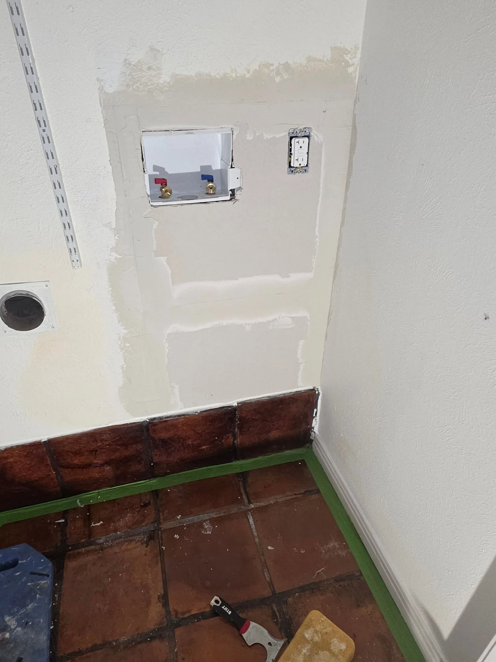 A laundry room corner with exposed drywall, a washer box, and a single electrical outlet above brown floor tiles.