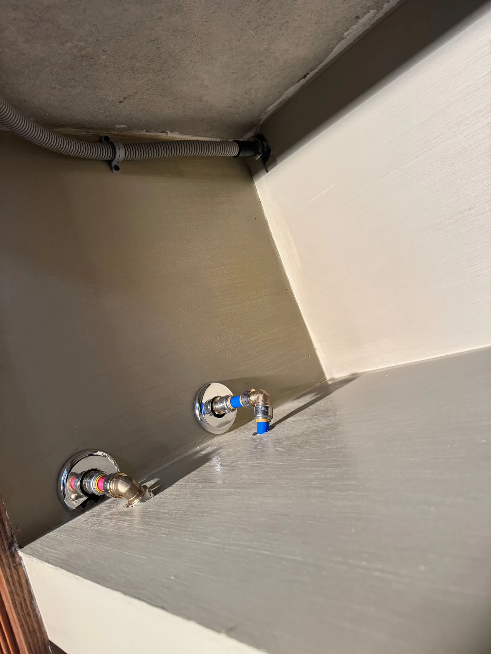 Inside of a kitchen sink cabinet with two new PEX pipe connections for hot and cold water supply, connected by chrome escutch