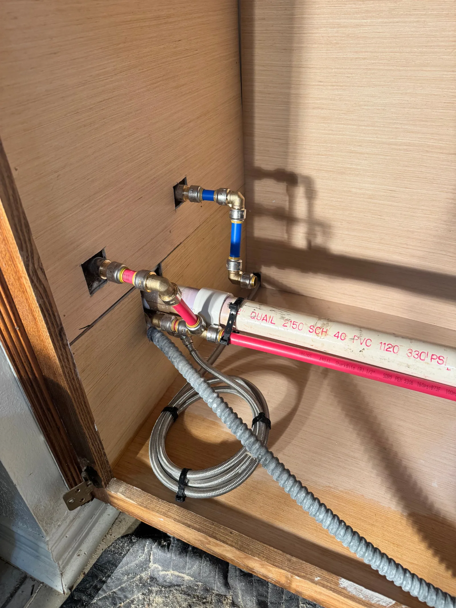 Pipes and plumbing connections in a light wood cabinet base. A white PVC pipe runs horizontally with red and blue PEX lines c