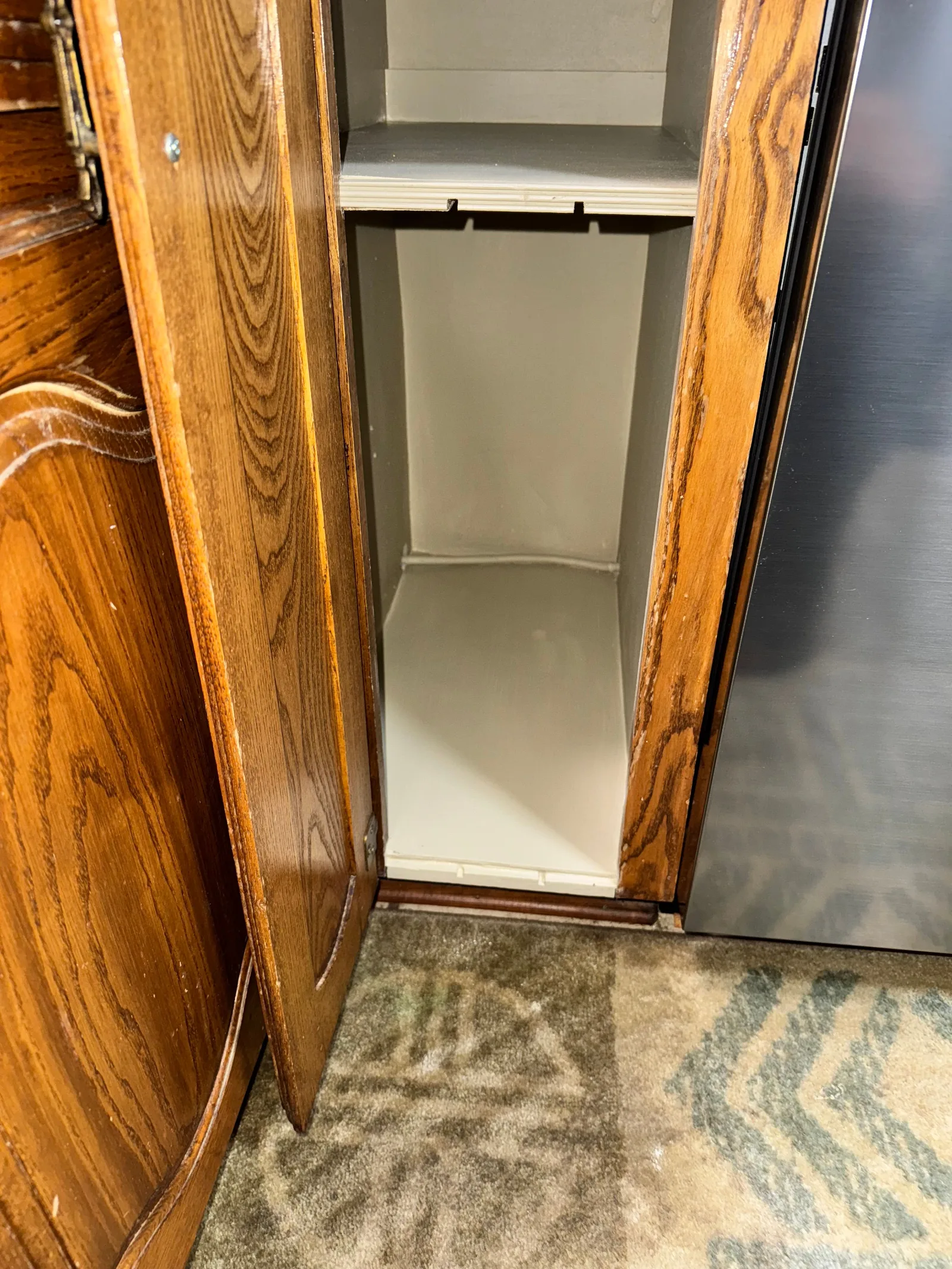 An open oak cabinet with a clean, light-colored painted interior, adjacent to a stainless steel appliance, above a carpeted f