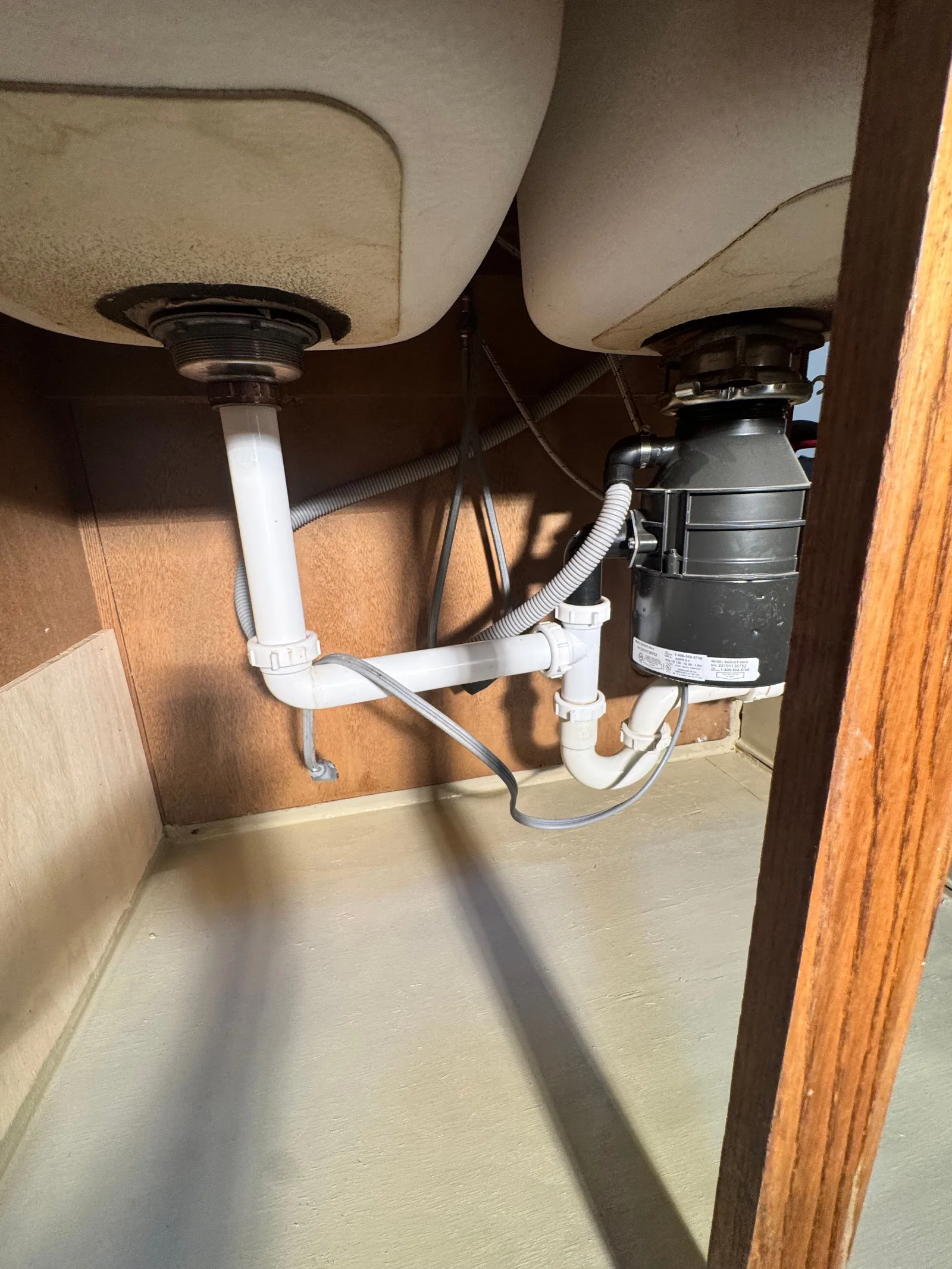 Under a kitchen sink, a dual white basin setup with an attached black garbage disposal unit, P-traps, and plumbing.