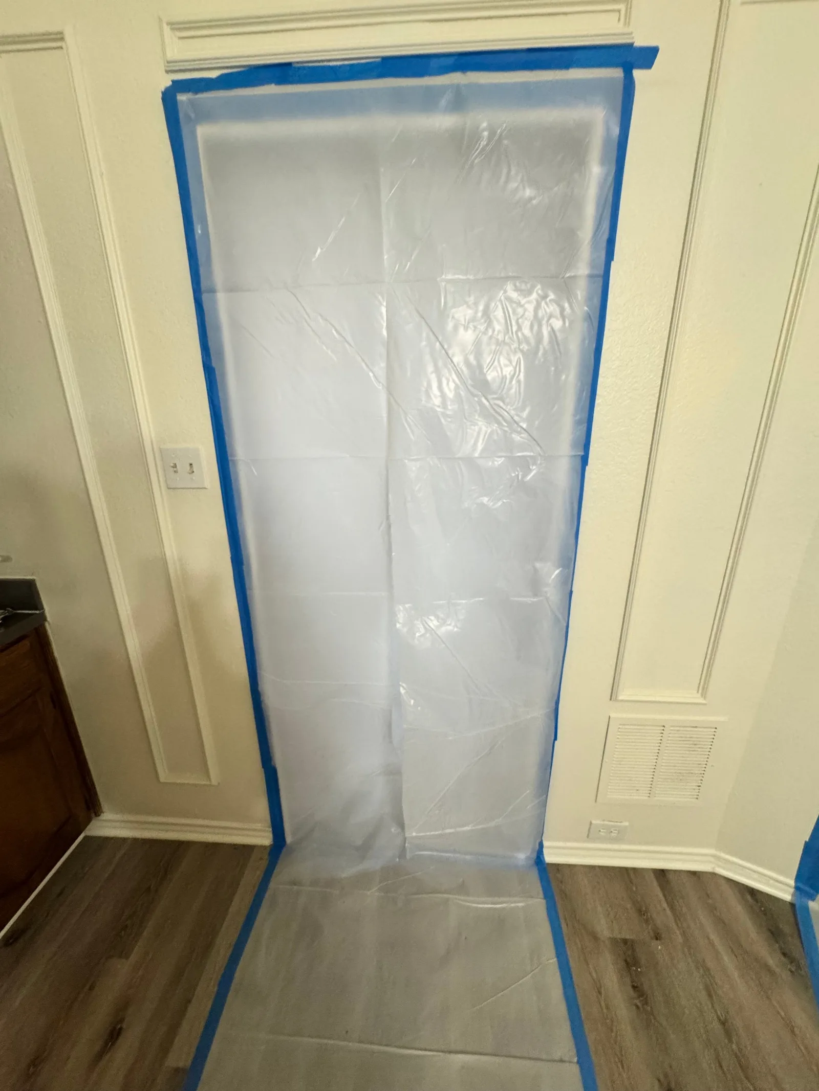 Plastic sheeting taped to a white molding doorframe and white textured wall over a wood-look floor, with power outlets visibl