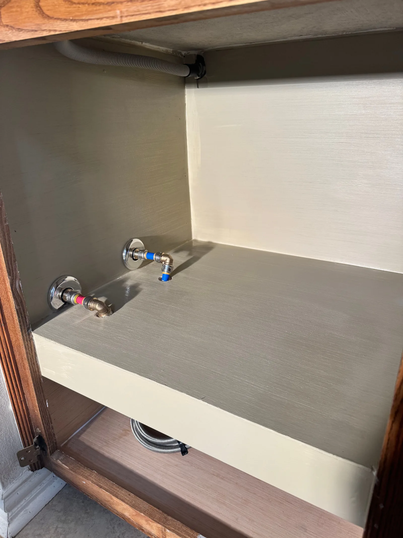 Interior of a newly cleaned and painted under-sink kitchen cabinet with new chrome plumbing fixtures and a corrugated pipe.