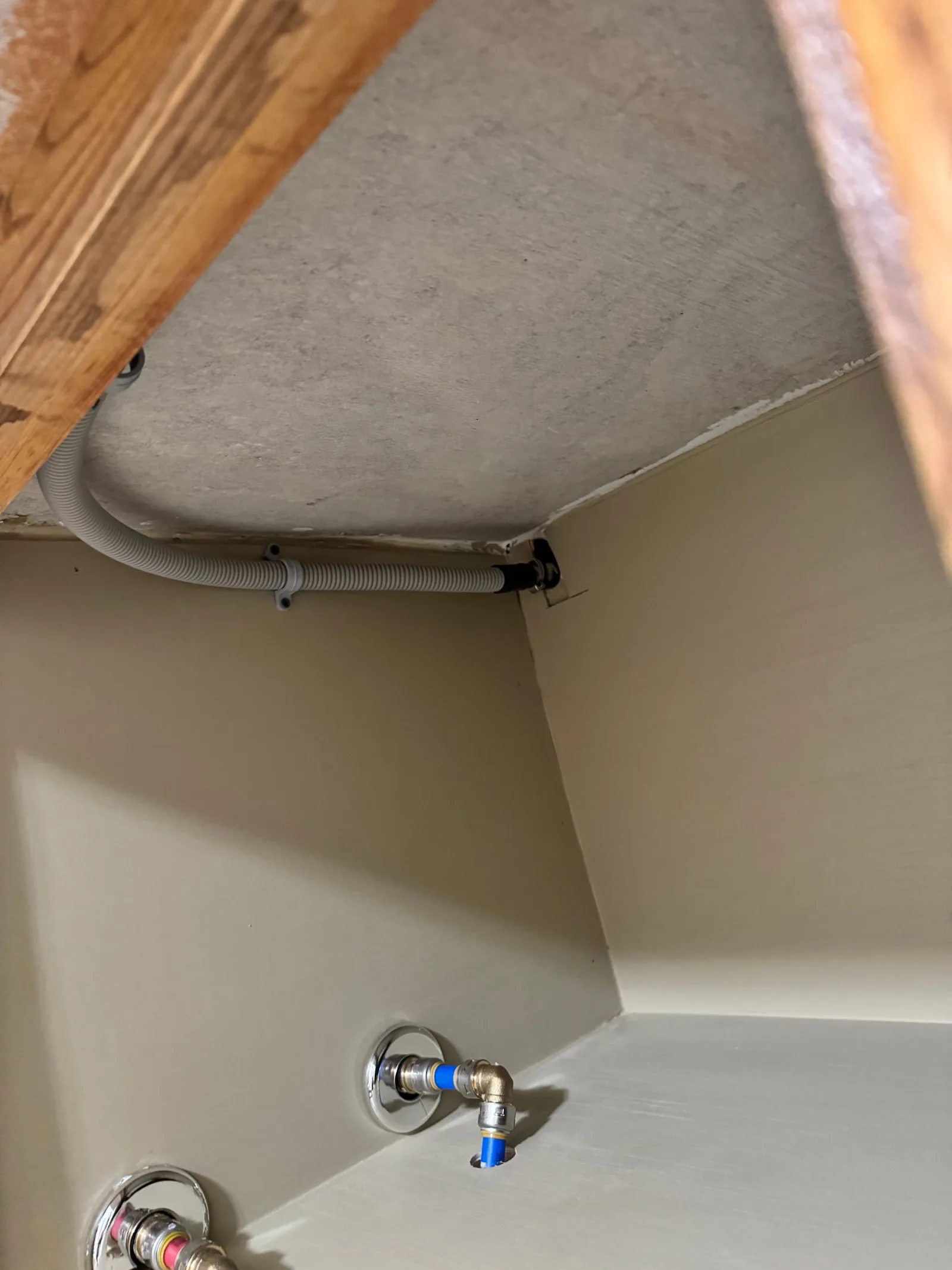 Underneath a wooden cabinet, a beige wall with a grey corrugated drain pipe curves, leading into a dark corner. Two PEX plumb