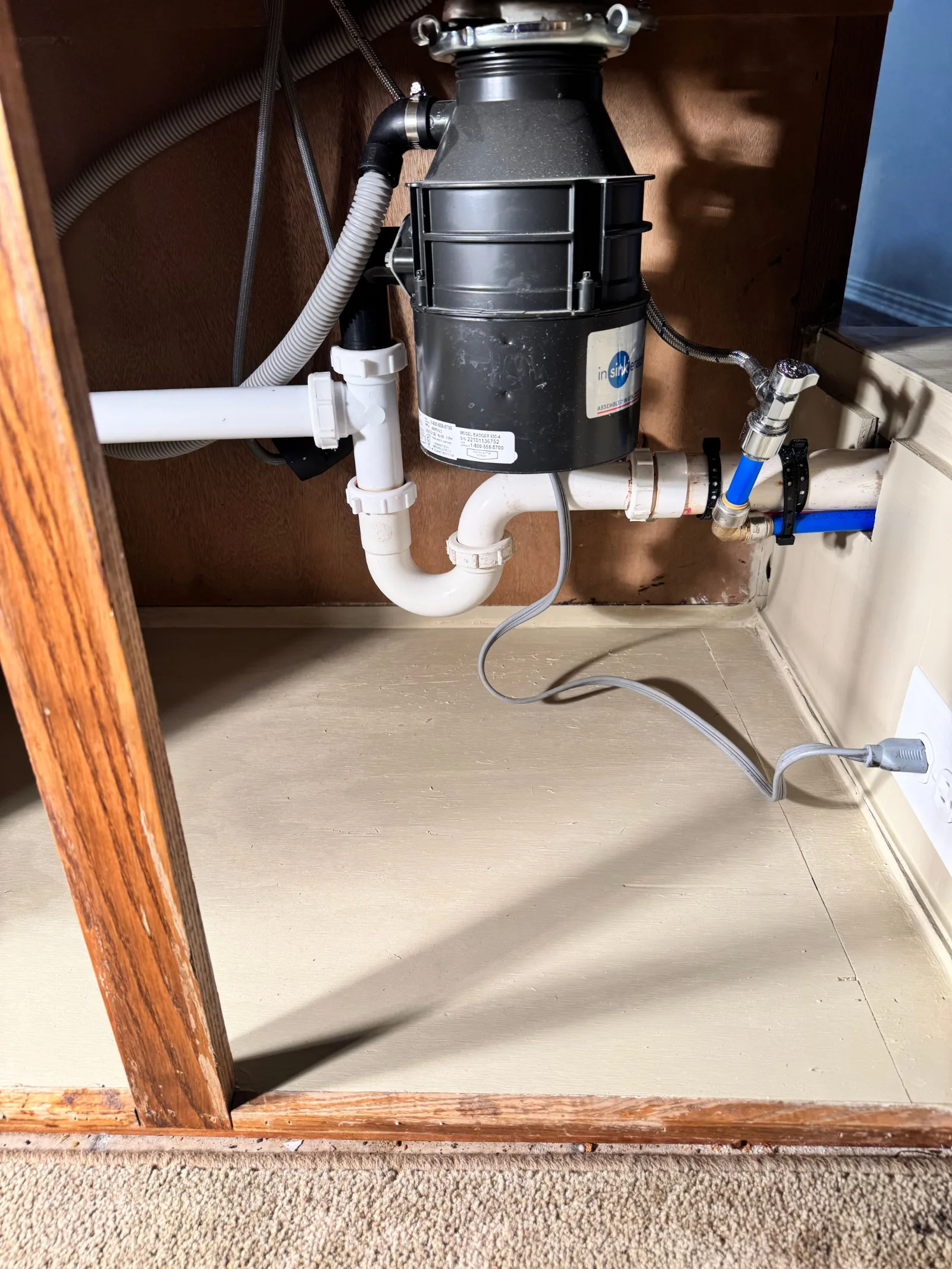 Under-sink plumbing with a black garbage disposal, white P-trap, blue PEX water line, and a gray power cord.